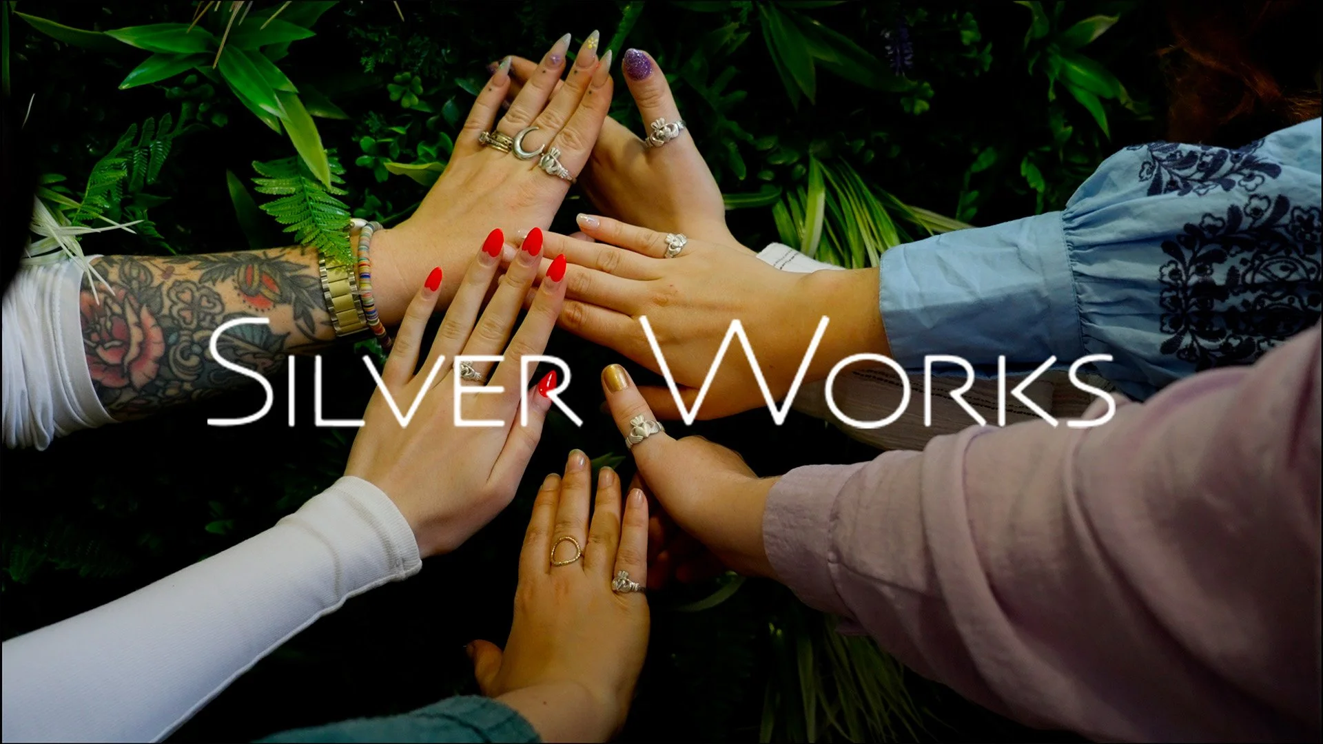 Multiple hands with various jewelry and nail polish placed on top of each other over green plants, with the text 'SILVER WORKS' overlaying the image.