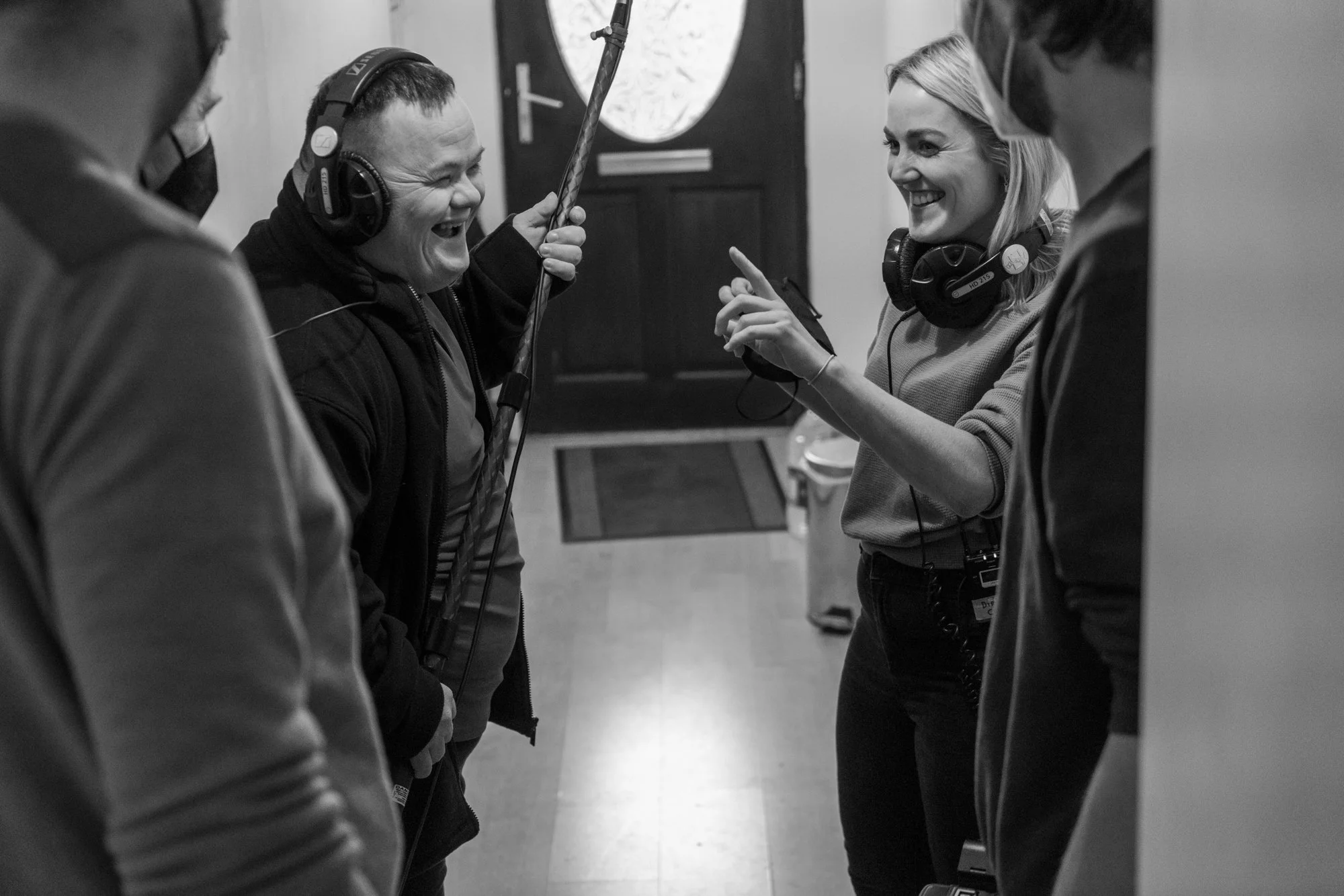Behind the scenes photo. Four people in a circle wearing headphones, engaging in a lively conversation or activity, in a room with a door and a window in the background.