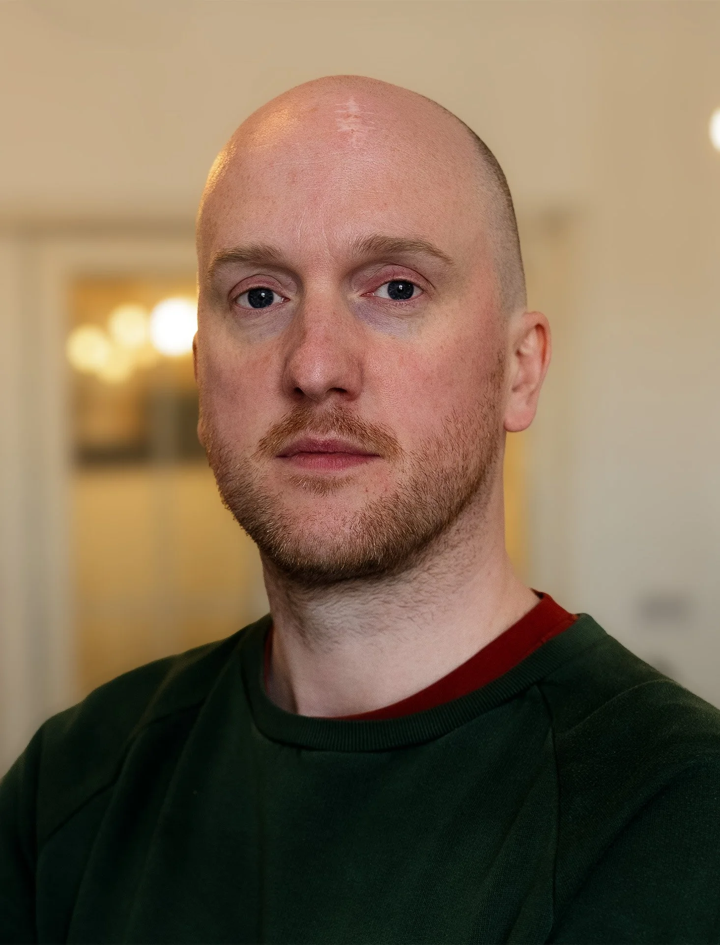 Close-up of a young man with a shaved head, light stubble, and light-colored eyes, wearing a dark green and red shirt, looking into the camera with a neutral expression, with a blurred background of indoor lighting.