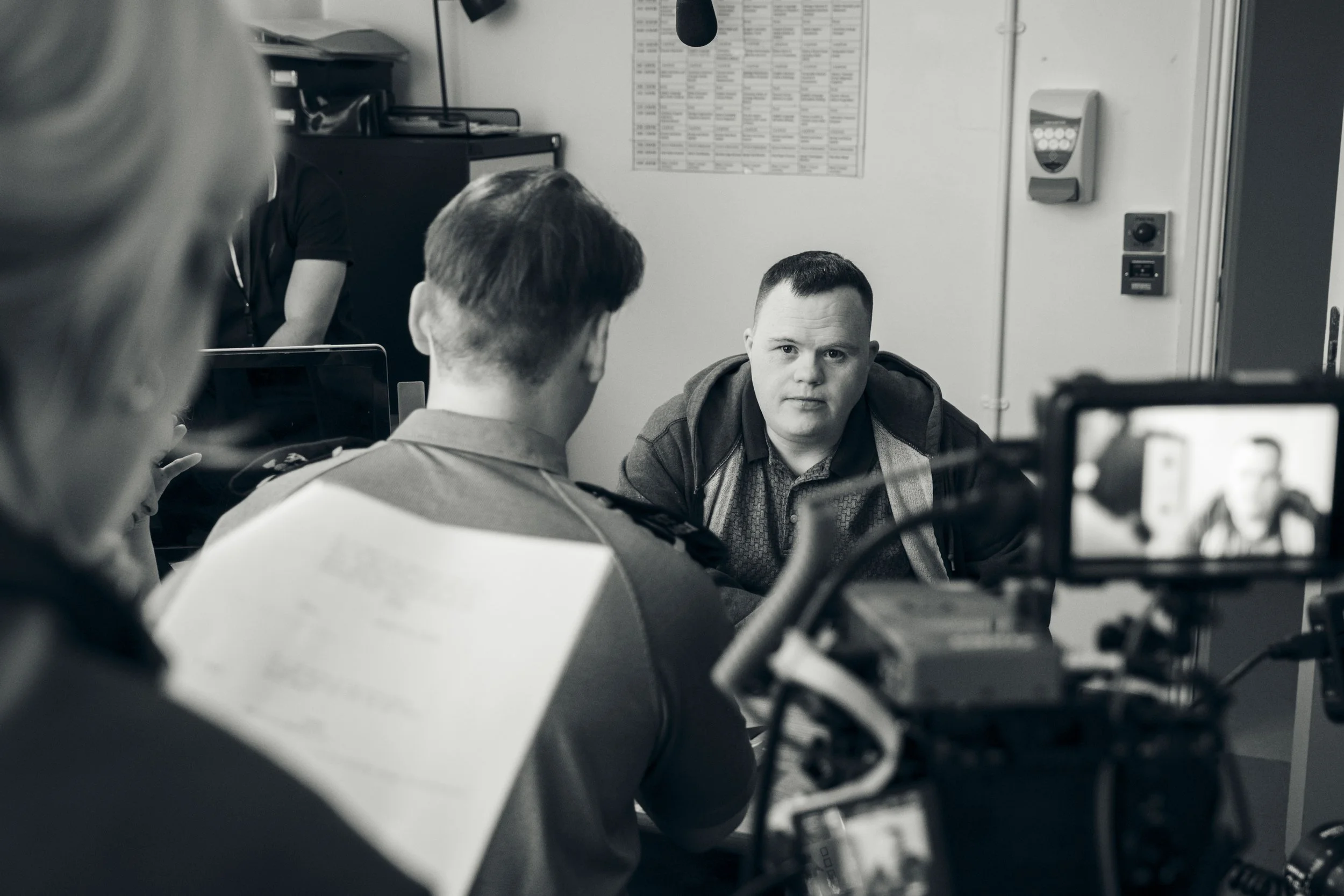 Black and white photo of a man sitting at a desk, facing another person whose back is to the camera. The man at the desk has a serious expression. There are various office objects, a computer, and a wall-mounted schedule or chart in the background.