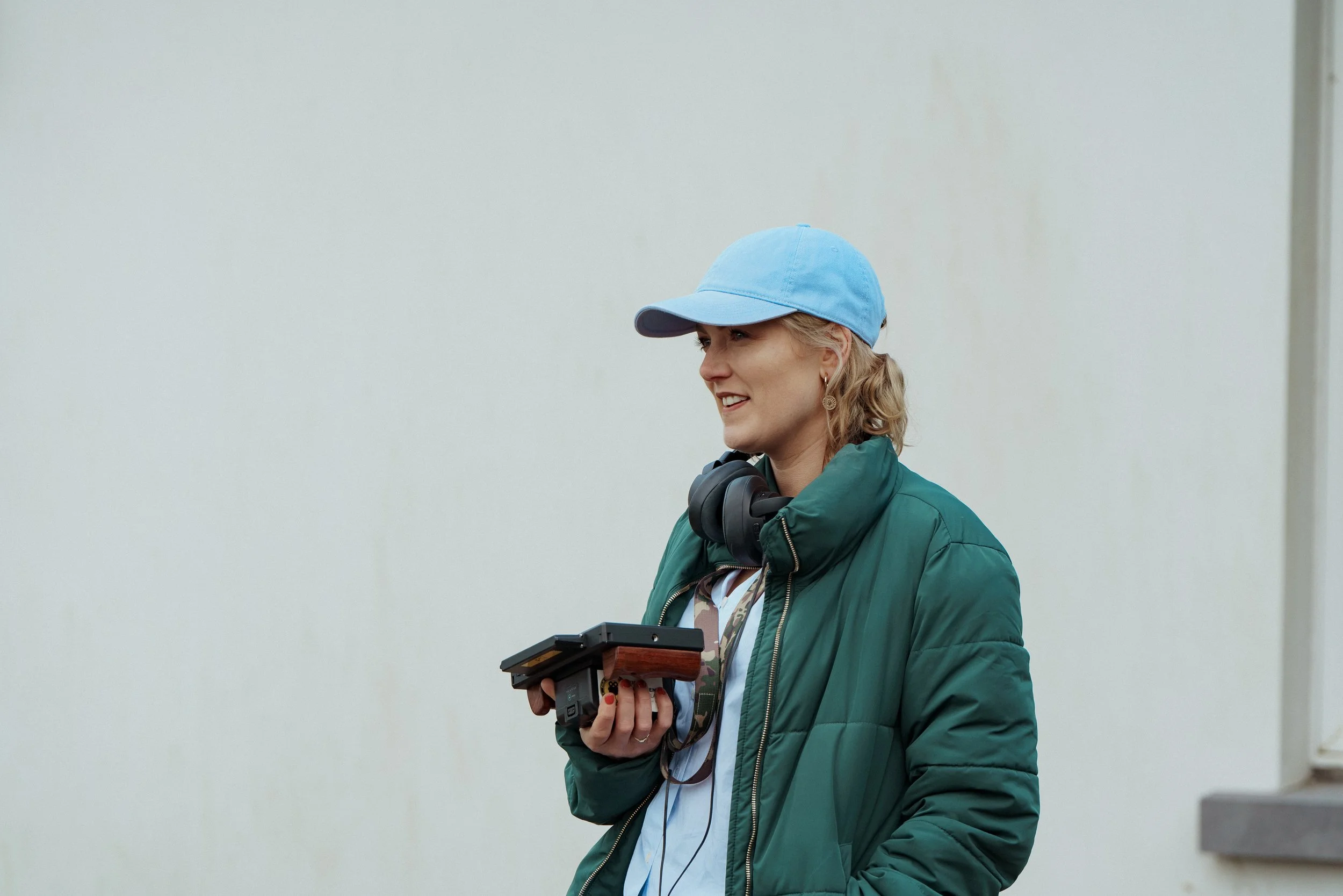 Woman with blonde hair, wearing a blue cap and green jacket, holding a recording device and wearing headphones around her neck, standing outdoors against a plain light-colored wall.