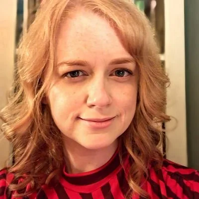 Close-up of a woman with curly strawberry blonde hair smiling, wearing a red and black striped shirt.