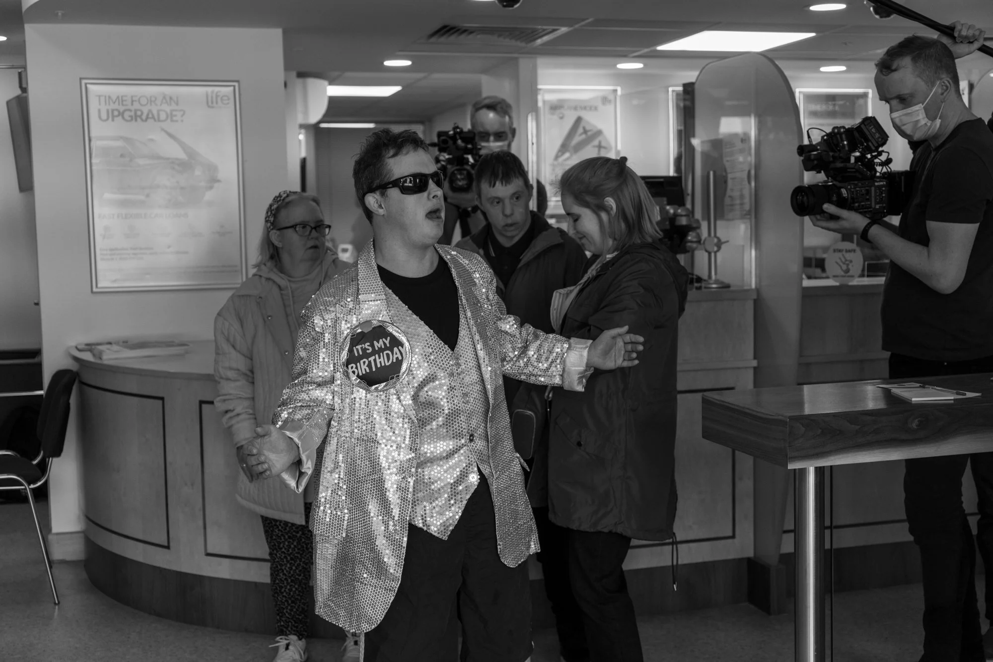 A person dressed in a sequined jacket with a birthday badge is being interviewed by a camera crew at a reception desk in a public indoor space, with several people gathered around and a poster advertising a product in the background.