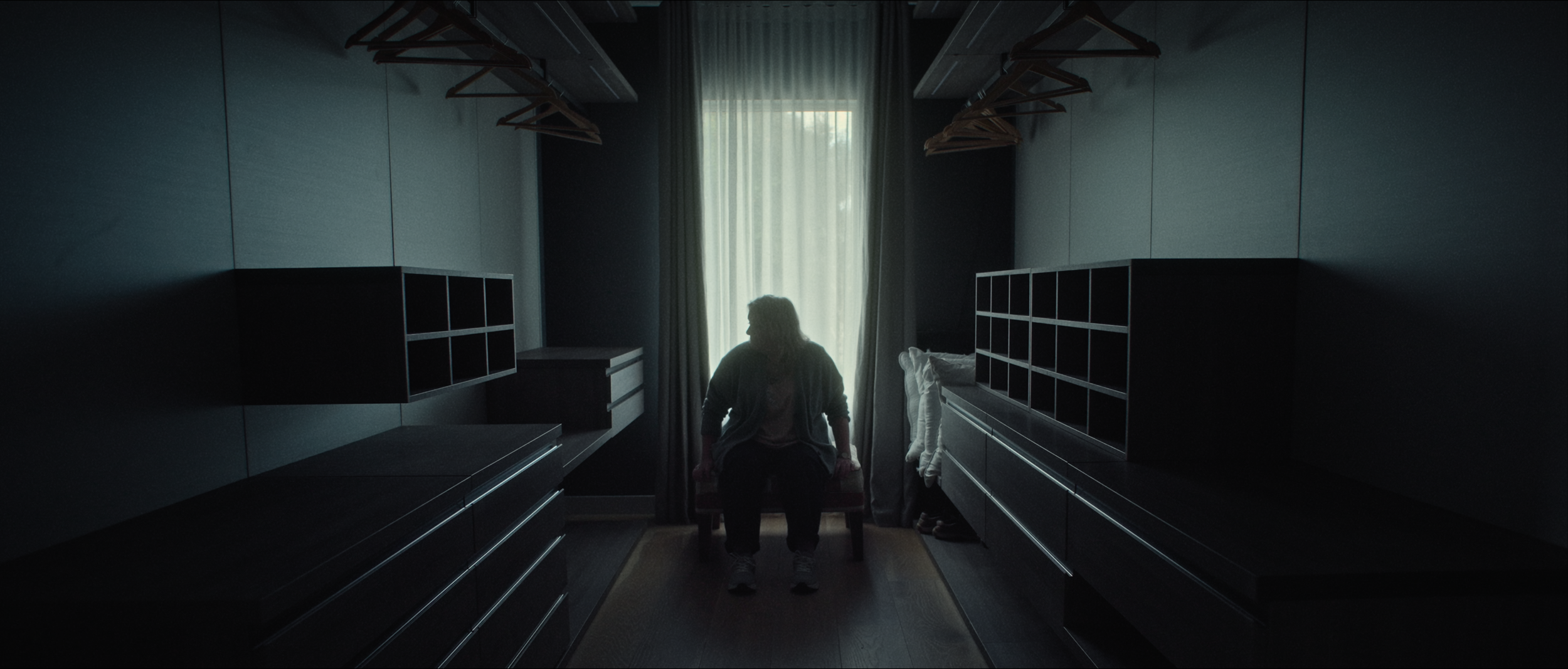 A silhouette of a person sitting on a bench in a dimly lit walk-in closet, facing a window with curtains, with empty shelves and hanging racks on either side.