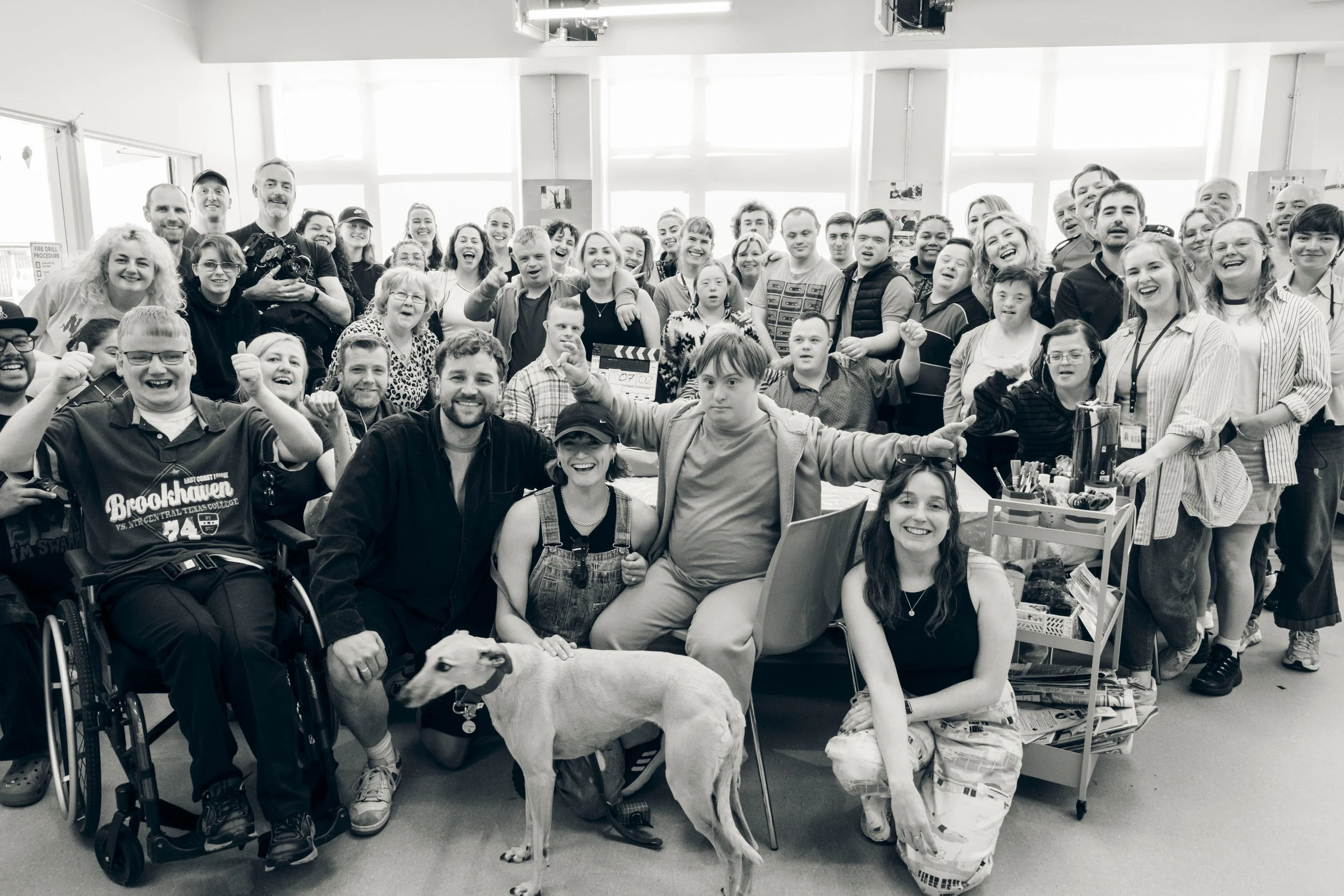 A large group of diverse people, including children, adults, and seniors, gathered indoors with big windows in the background. They are smiling, laughing, and posing for the photo. Some are making gestures like thumbs up. There is a dog in the foregr