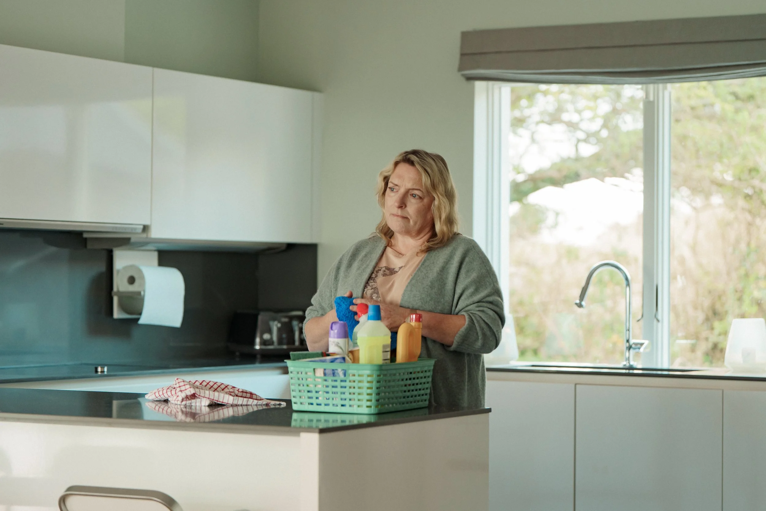 A woman with blonde hair standing in a modern kitchen, looking away with a concerned expression, at a countertop with a green basket of cleaning supplies, a dish towel, and a window in the background.