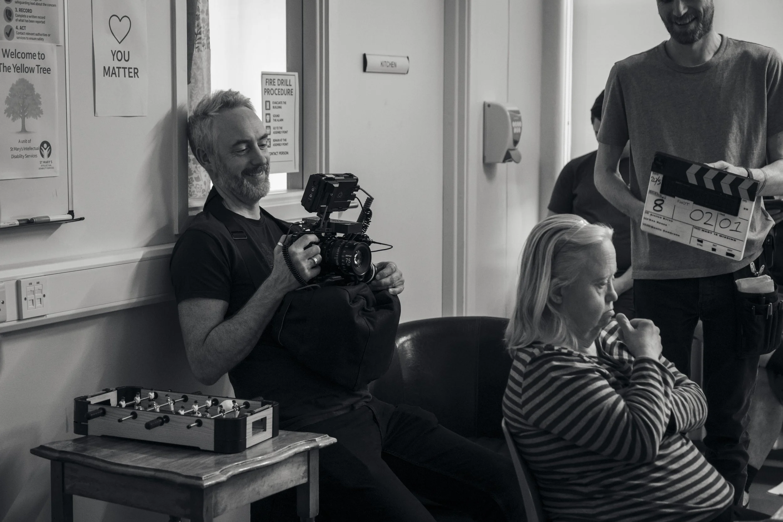 A group of people, including a man sitting on the floor with a camera and another woman sitting on a chair, in a room with posters on the wall. One person is holding a clapperboard. The scene appears to be a filming or video production set.
