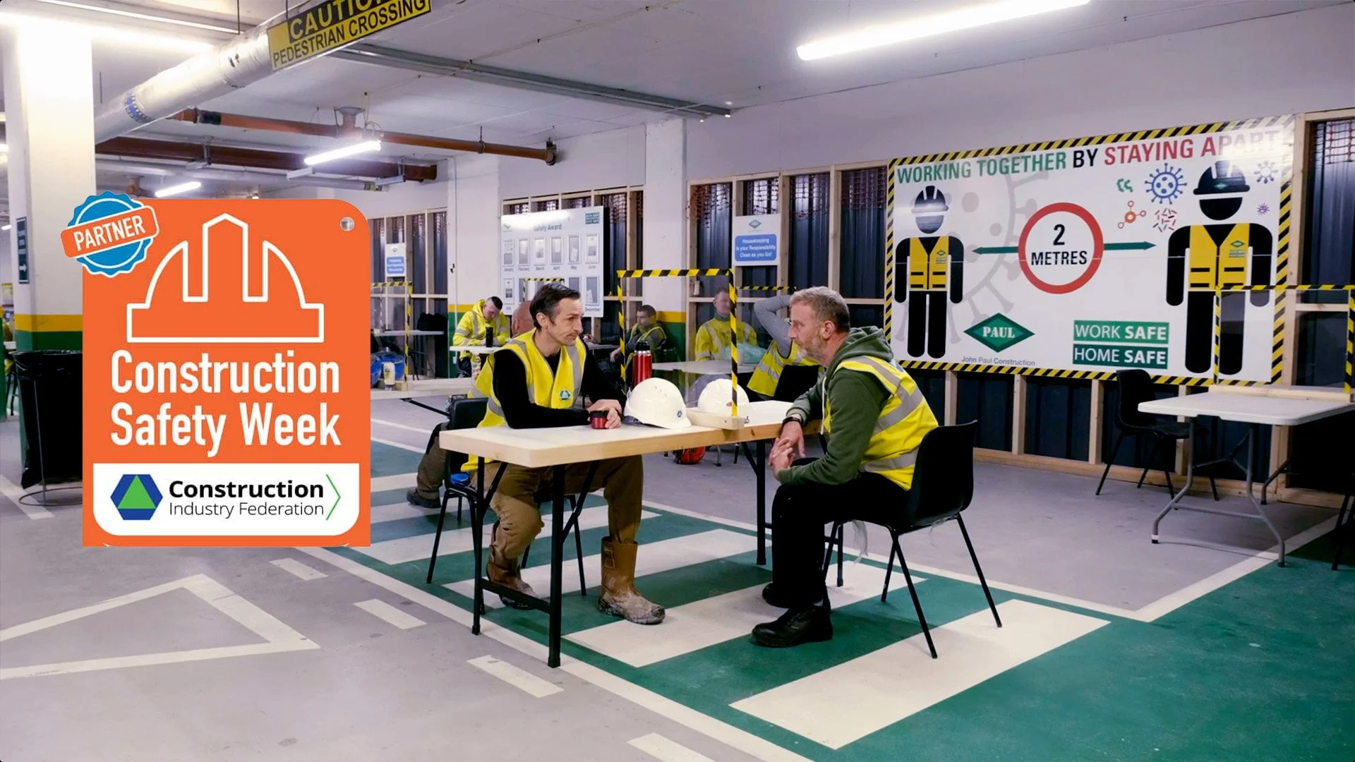 Construction workers sitting at a table in an indoor training area, with safety signs and posters on the walls, including a large banner about keeping 2 meters apart for safety.