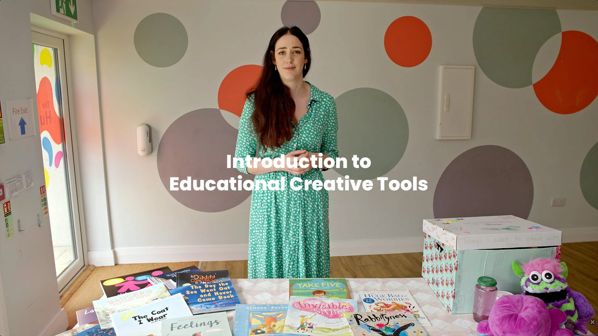 A woman standing in a colorful room with books and toys on a table in front of her, and a box labeled 'Things I want to share with you.' The room has a white wall with large polka dots, a door with colorful designs, and a fire exit sign.