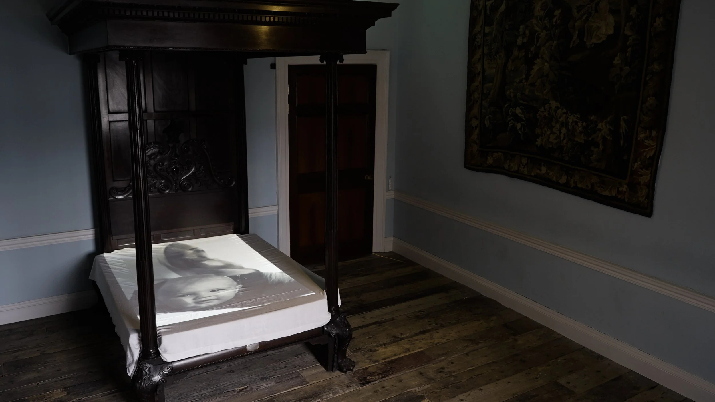 A four-poster bed with an ornate dark wood headboard, with a duvet featuring black-and-white images of a child's face and a person's face. The room has light blue walls, dark wooden flooring, and a tapestry hanging on the right wall.