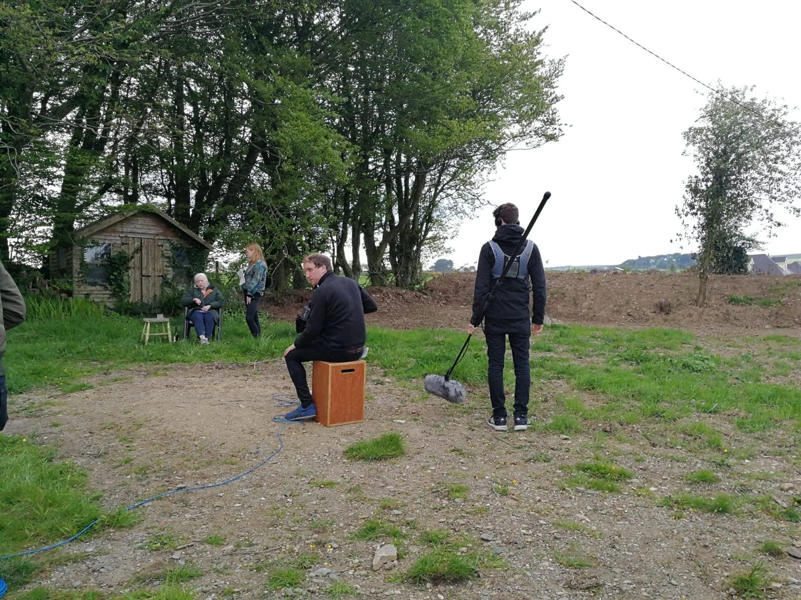 A film crew filming outdoors in a grassy area with trees and a small shed in the background. Multiple people are engaged in different activities, including a seated elderly woman, a young woman standing nearby, and a man sitting on a wooden box with 