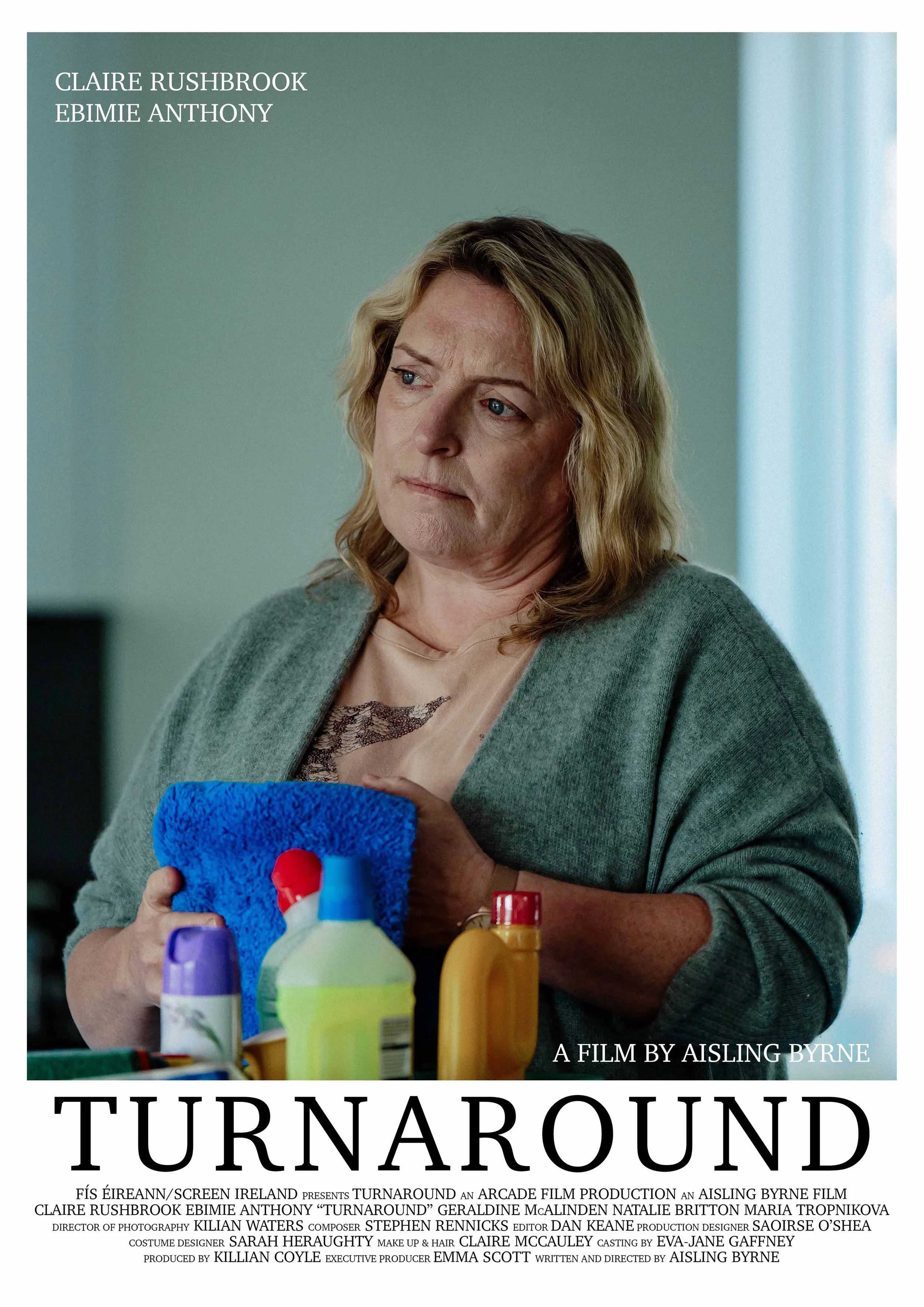 A woman with shoulder-length blonde hair, wearing a gray cardigan over a beige top, holding a blue cleaning cloth and looking concerned, standing behind a table with various cleaning supplies. The background is a softly lit room with a window.