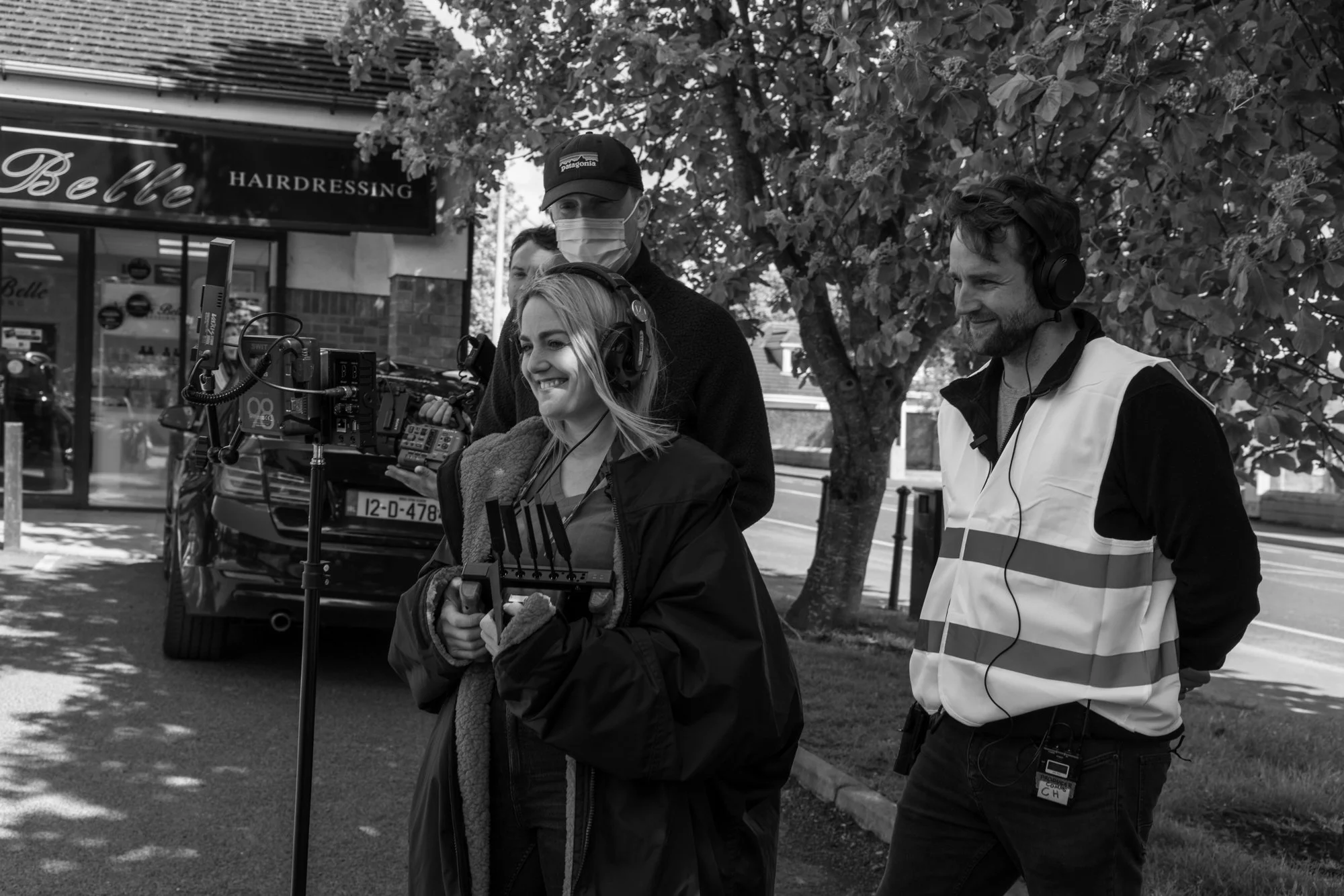 Crew filming a scene outdoors, with a woman operating a camera rig, a man wearing headphones and a vest, and another person wearing a mask and a Patagonia cap, beside a tree and a hairdressing storefront.