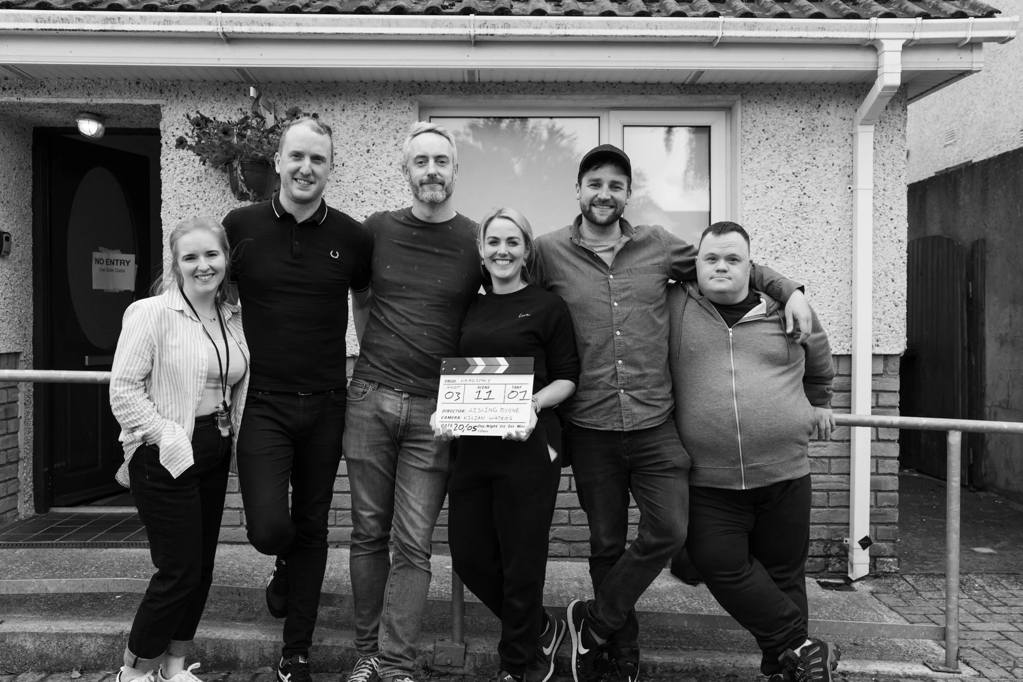 A group of six people standing in front of a house, smiling and posing for a photo during a film shoot, holding a clapperboard.