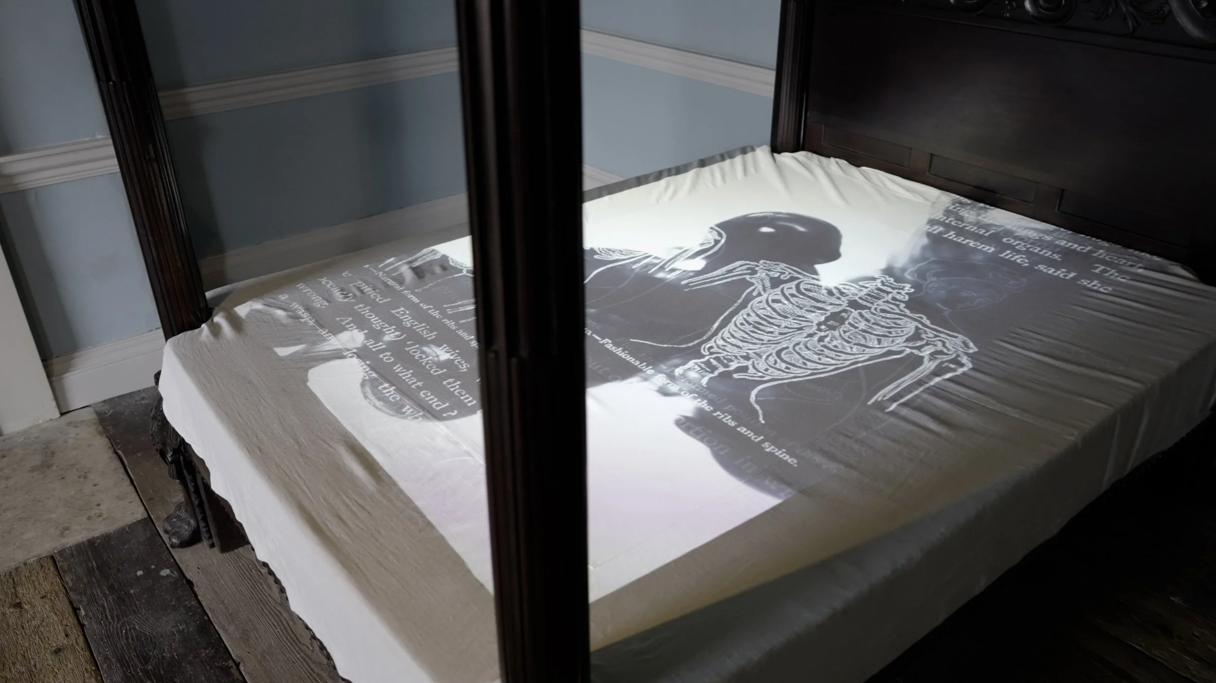 A bed with a projection of a skeleton and text on the white bedsheet, seen through glass panels.