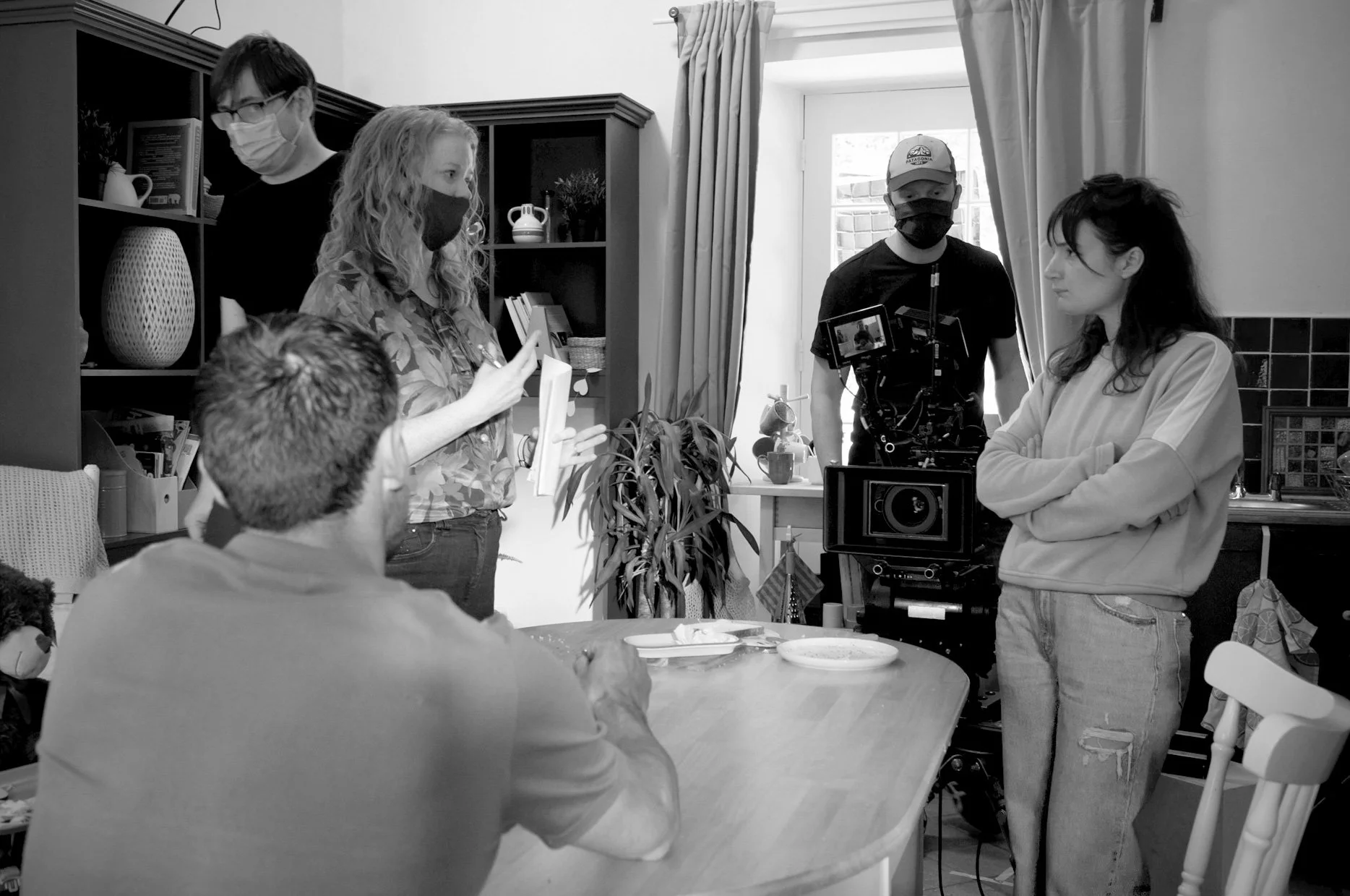 A group of people on a film set, including a woman with long curly hair and a camera operator, are gathered in a well-lit room with a window and curtains. Several of them are wearing masks. One woman is holding a piece of paper, and another person in