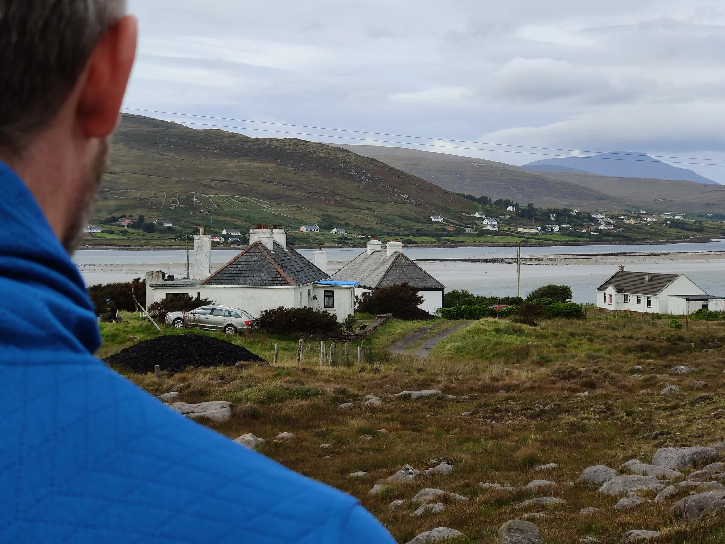 A person wearing a blue jacket is outdoors, overlooking a scenic view with rolling hills, scattered houses, and a body of water under a cloudy sky.
