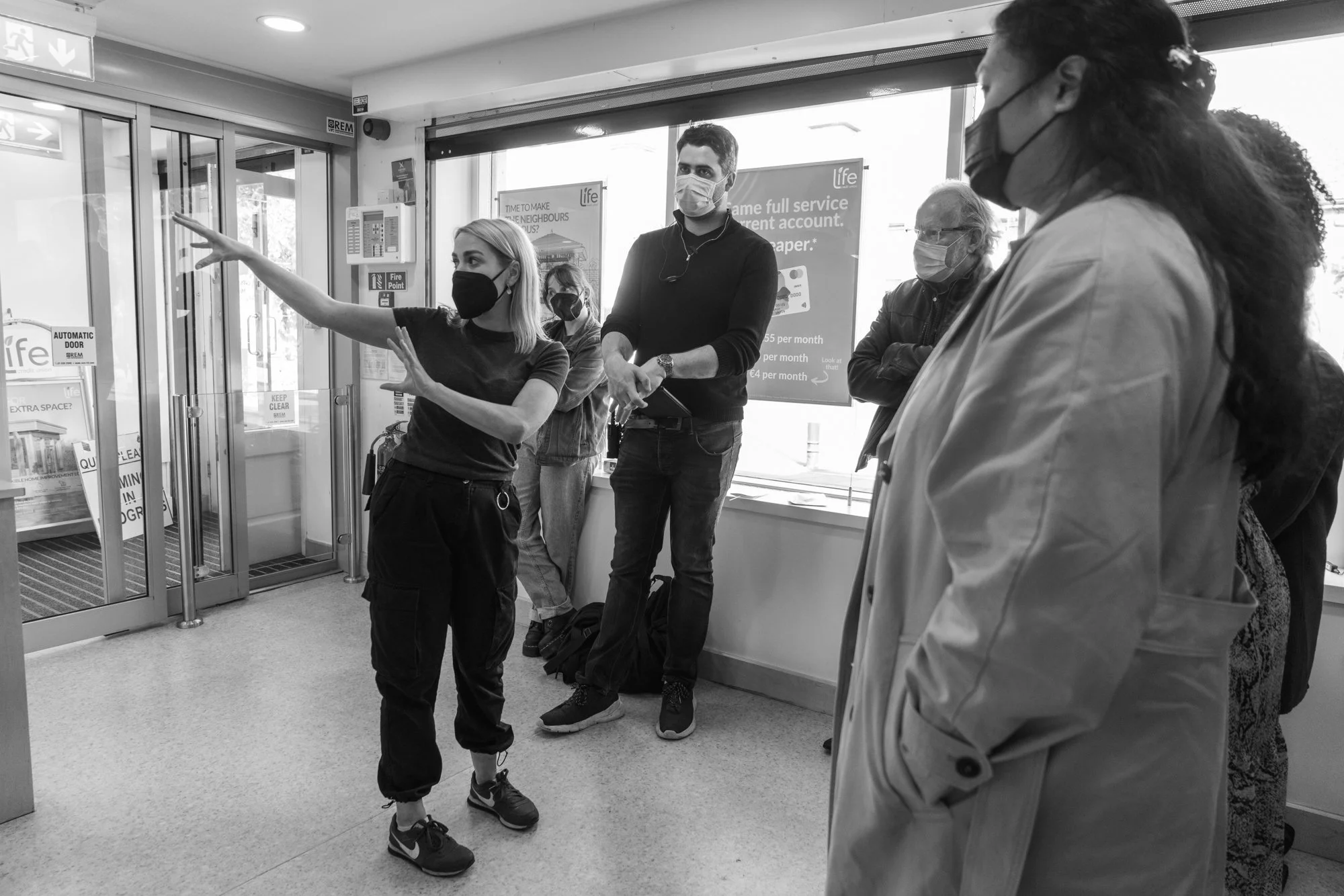 Group of five people wearing face masks inside a building near glass doors, with one woman pointing towards the doors.