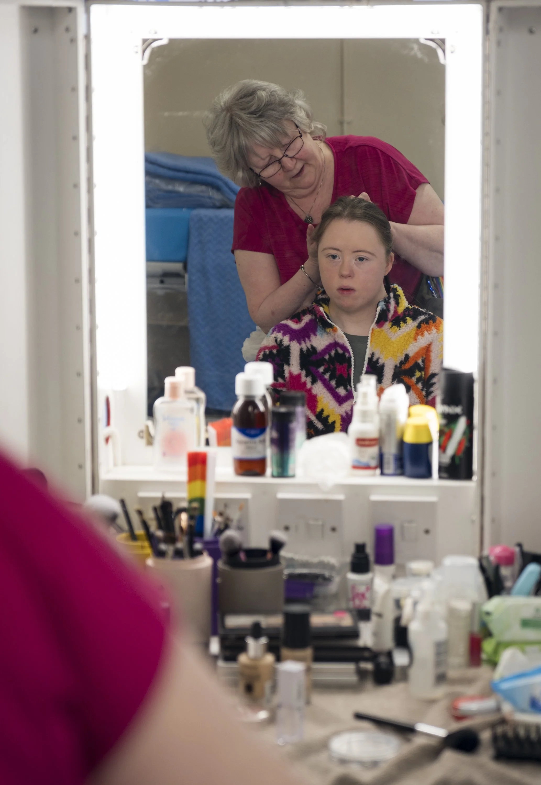 A mirror reflecting a woman with glasses and short gray hair doing a makeup or skincare routine on a young girl with brown hair wearing a colorful patterned hoodie. The foreground has various bottles of makeup and skincare products on a table.