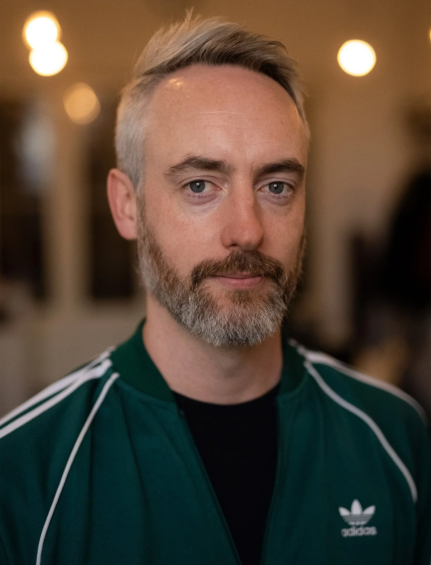 Close-up of a middle-aged man with gray hair and a beard, wearing a green Adidas sports jacket with white stripes, in an indoor setting with warm lighting.