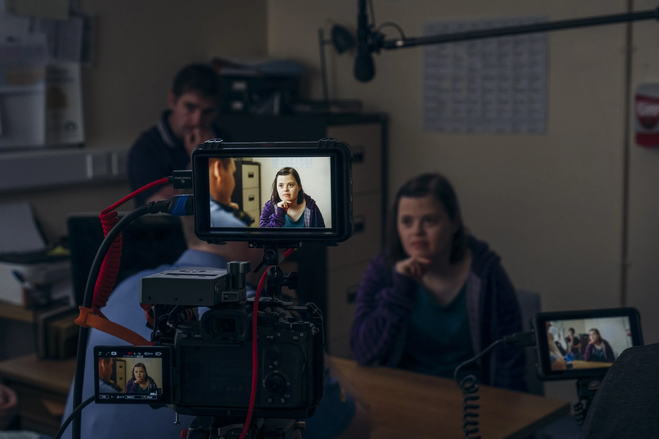 Behind the scenes of a video shoot showing a woman being filmed on a camera monitor, with two other women in the background engaged in conversation.
