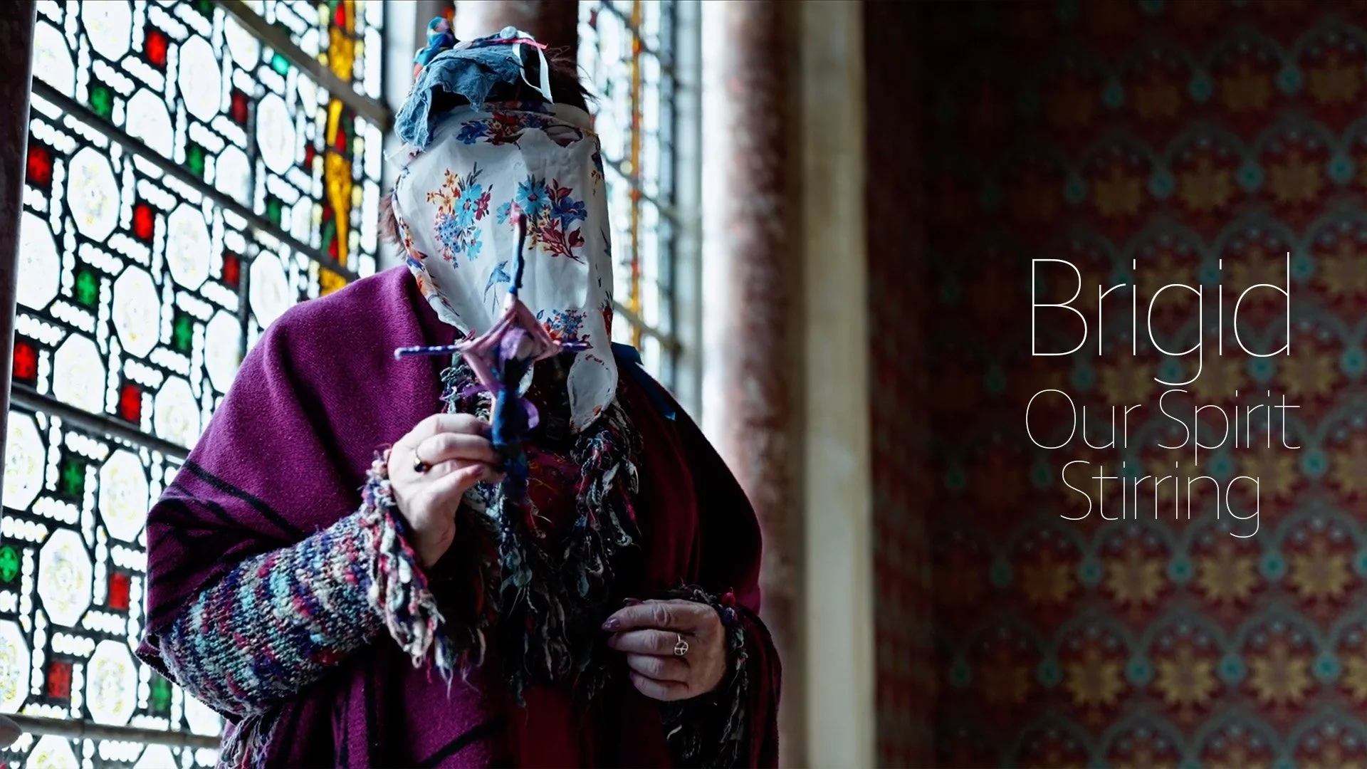 A person wearing a headscarf and a maroon shawl standing near stained glass windows, holding a handmade doll, with the text 'Brigid Our Spirit Stirring' overlayed.