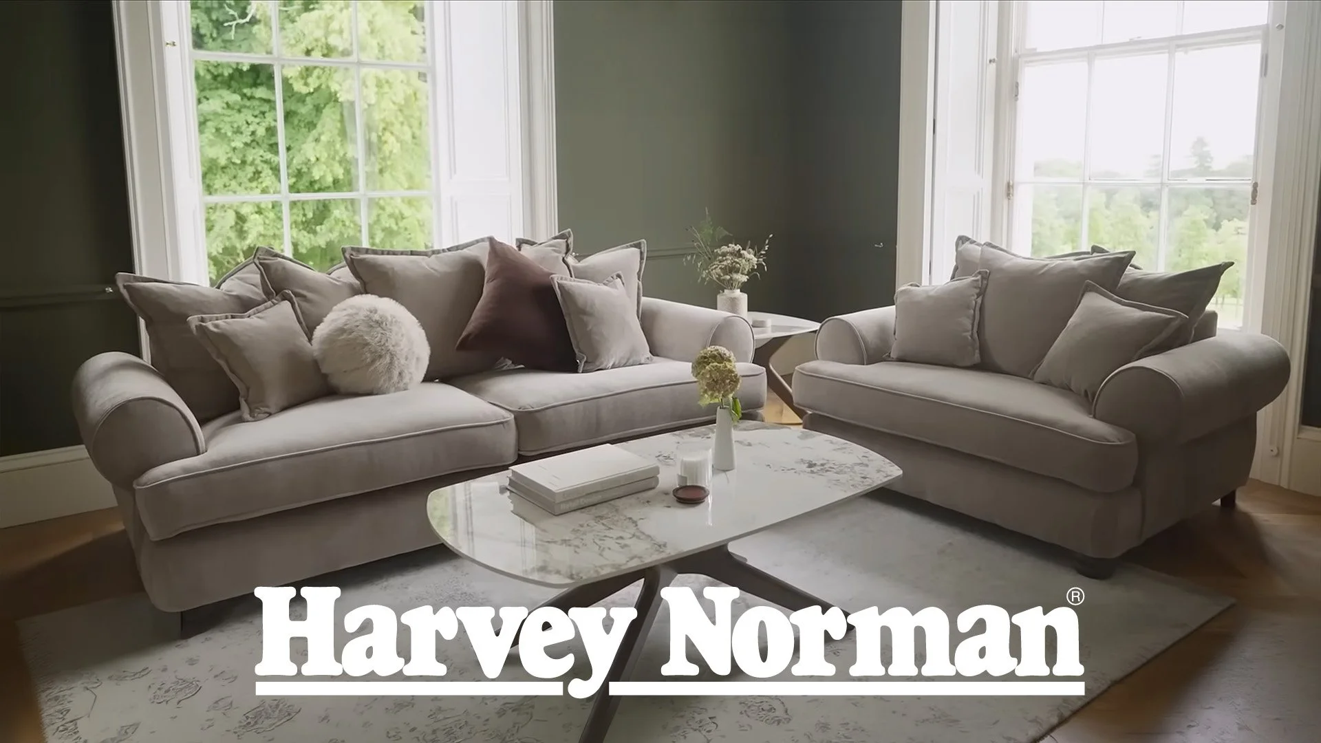 Living room with two beige sofas, coffee table with books and flower vase, large windows with green trees outside, neutral decor.