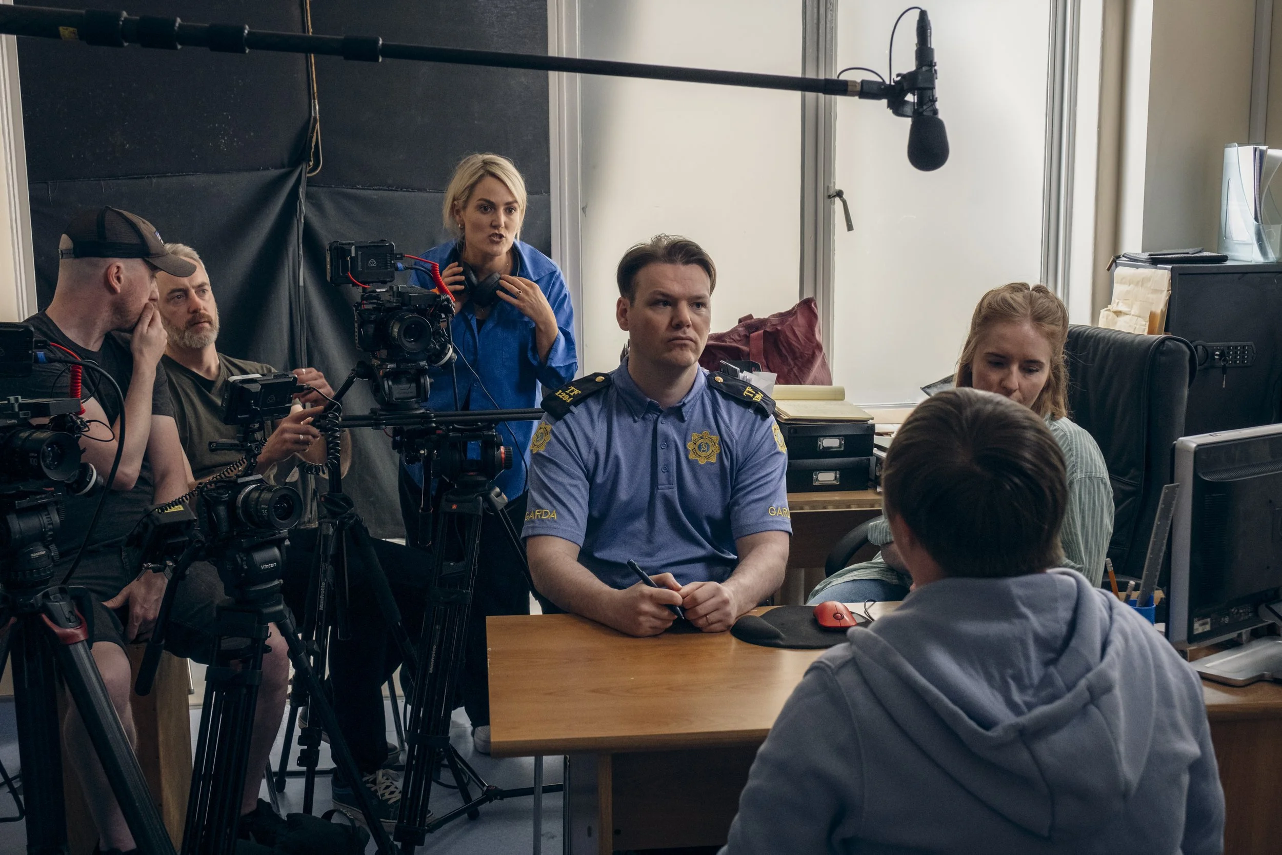 Group of people in a room with a camera and sound equipment, some seated at a table and one woman speaking, possibly filming a interview or documentary.