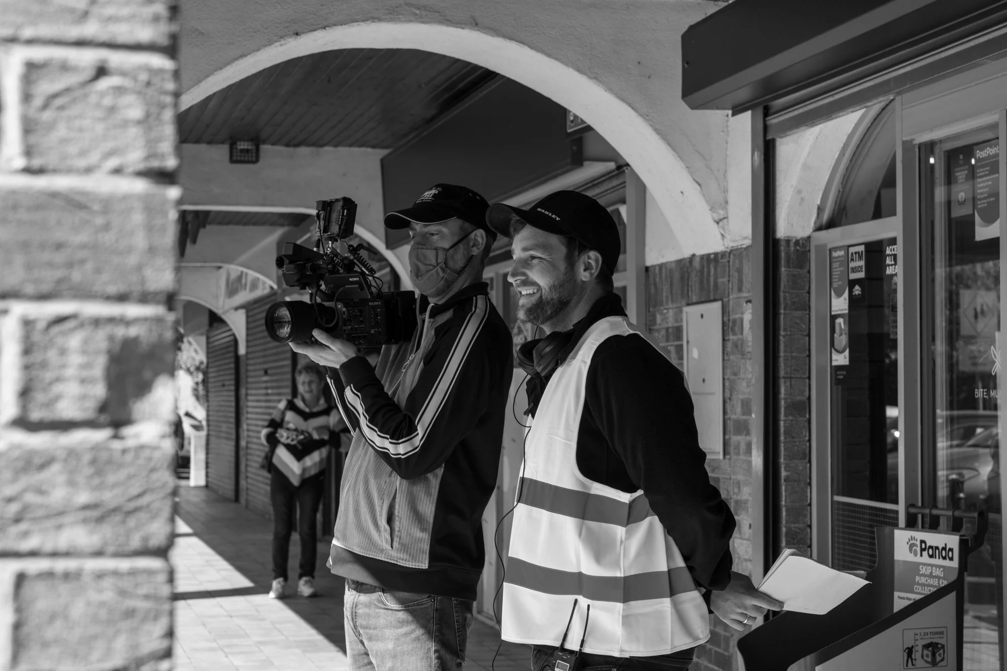 Two men, one wearing a mask and the other a cap with earphones, look at a camera while standing outside a storefront. One man is wearing a high-visibility vest and holding a folder, the other is operating a professional video camera. There are people