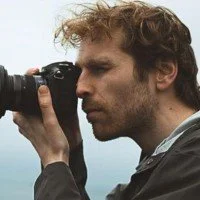 A man with curly hair and a beard looking through a camera's viewfinder, outdoors against a cloudy sky.