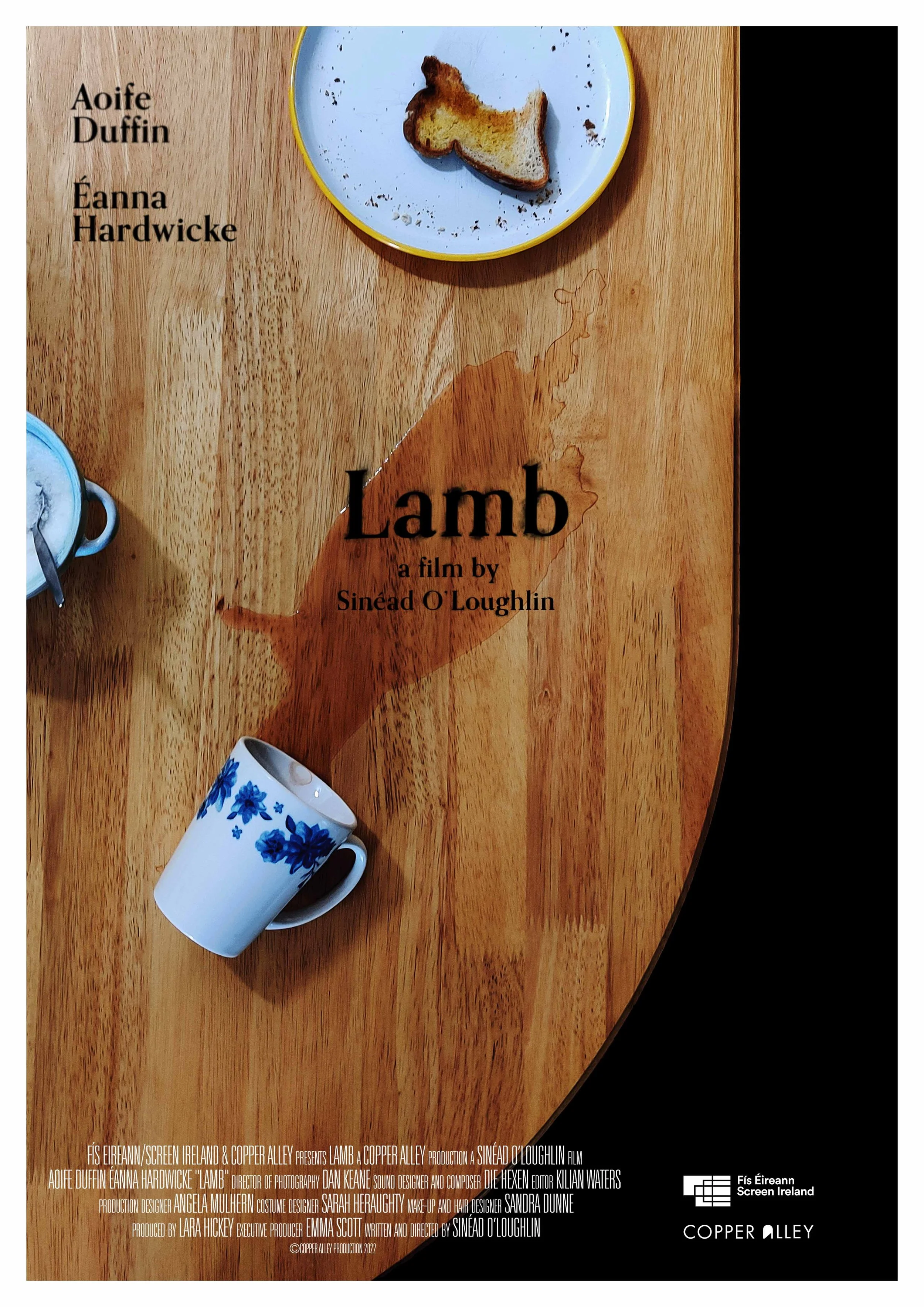 A wooden table with a blue plate holding a piece of toast, a white mug with blue flowers, and another mug with a spoon inside. Overlaid text reads 'Lamb, a film by Sinéad O'Loughlin,' with credits at the bottom.