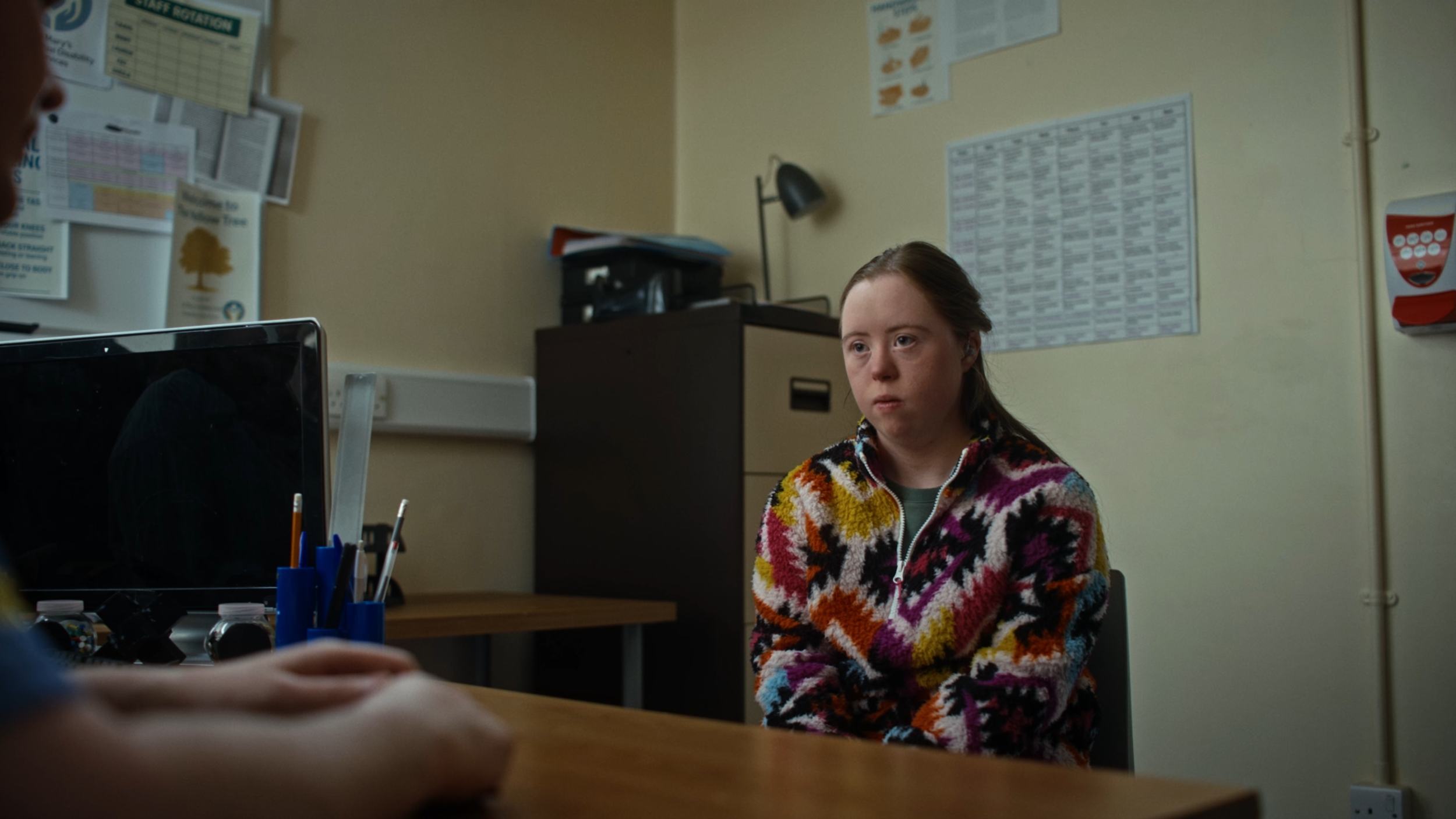 A young girl with light skin and brown hair sitting in an office or clinical setting, wearing a colorful patterned fleece jacket, looking serious or concerned. There are posters and charts on the wall behind her, a desk with a computer, and a person’s arm partially visible in the foreground.