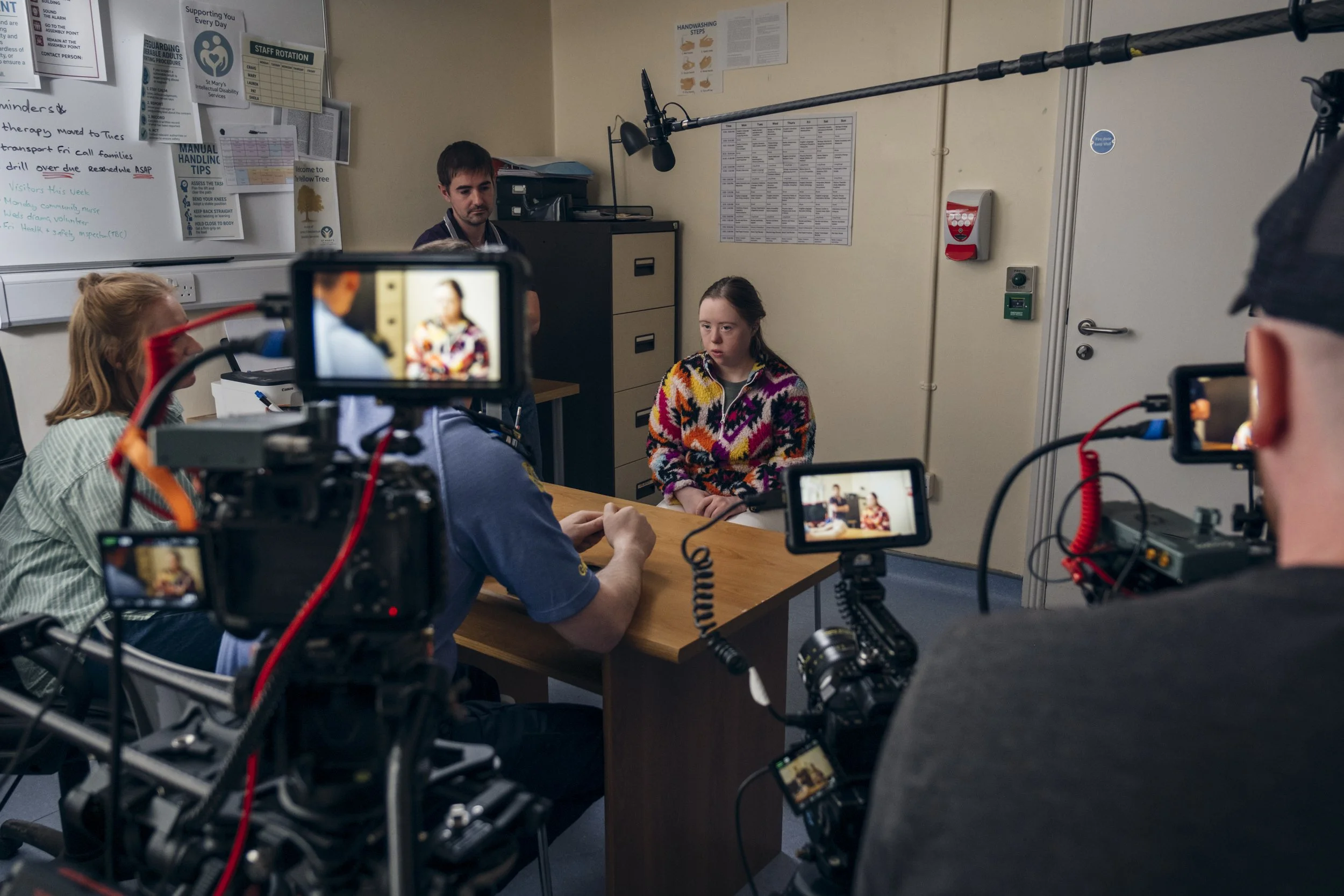 A young girl with brown hair and a colorful sweater sits at a table with a man, while a camera crew records them in an indoor setting. The background features a bulletin board, posters, and a door.