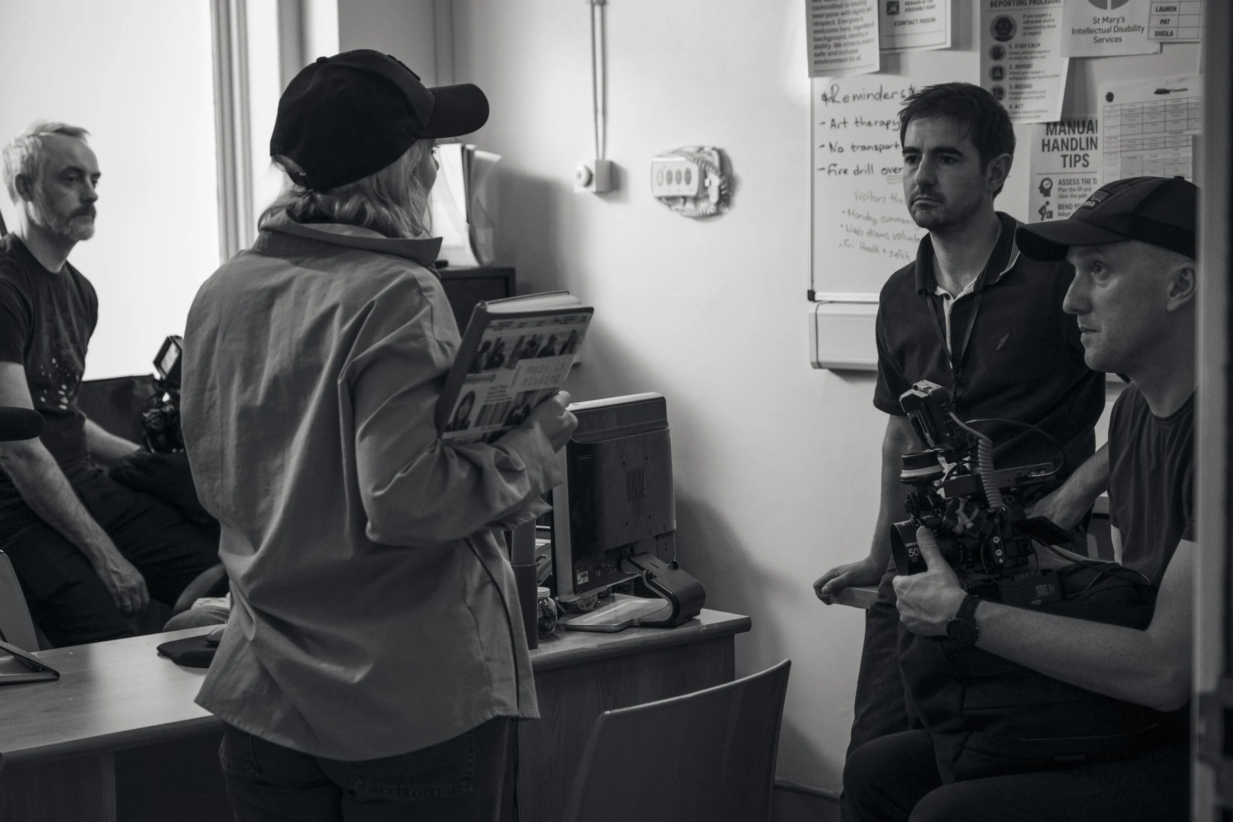 Four people in a room having a conversation. One woman with a cap and a coat holds a folder, while three men, two wearing caps, are listening. There are posters and notes on the wall behind them.