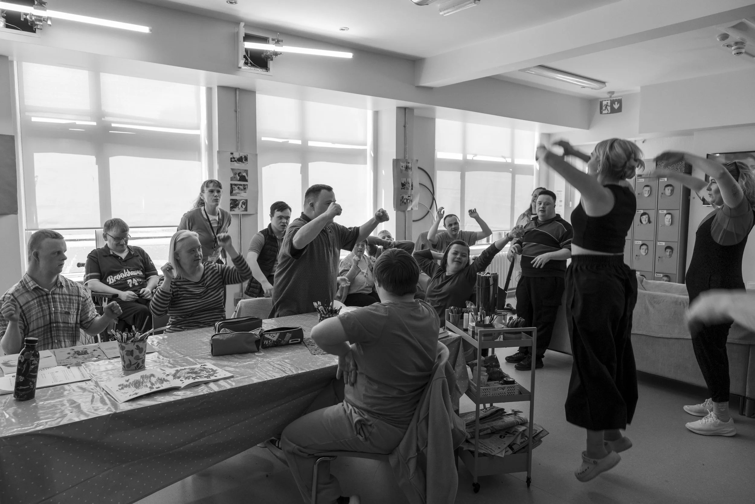 A group of children and two adult women in a classroom engaged in a lively activity, with some raising their arms and others seated at a table covered with art supplies, in front of large windows.