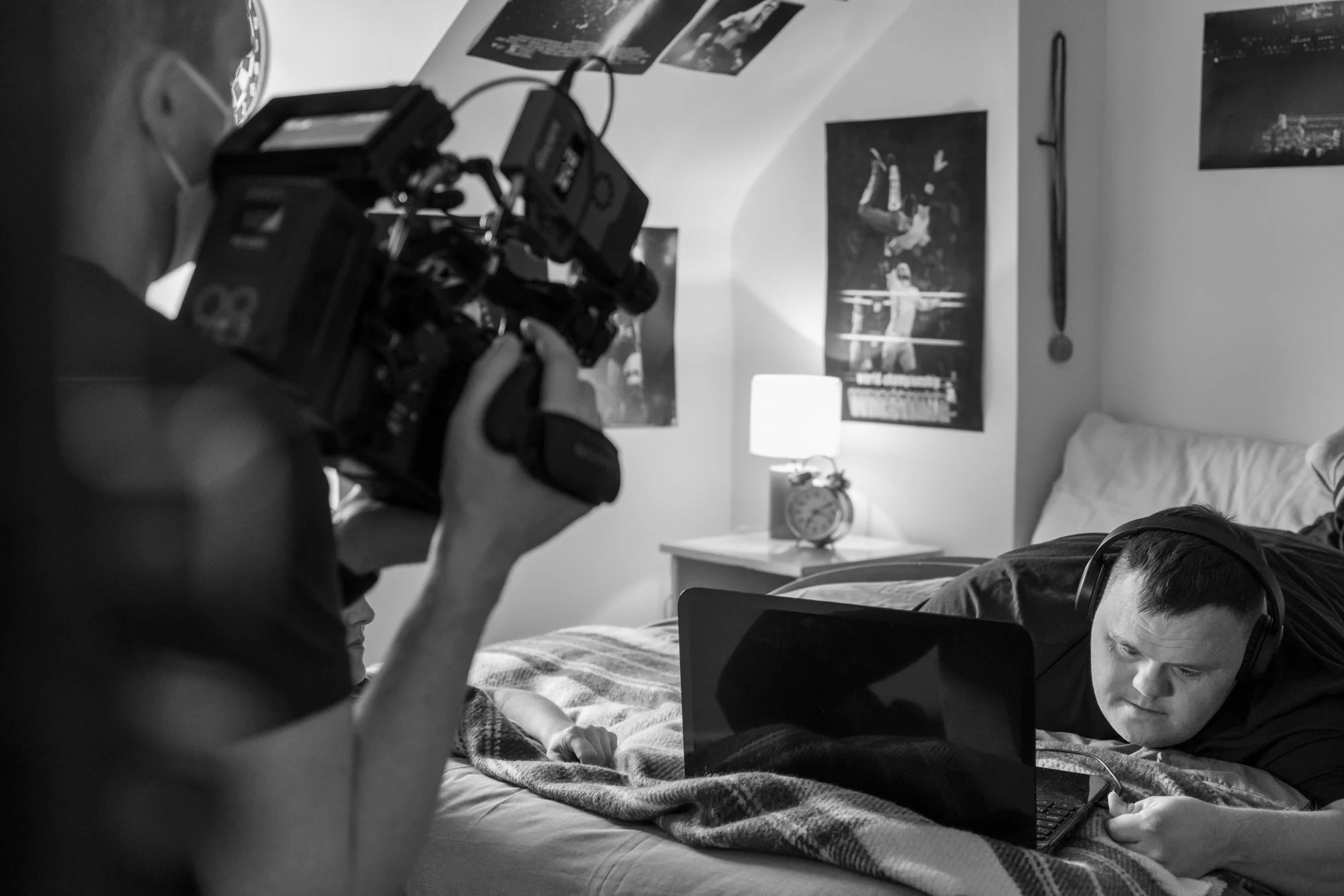 Black and white photo of two young men with headphones using laptops lying on a bed, with a person taking a photo or video of them with a camera.