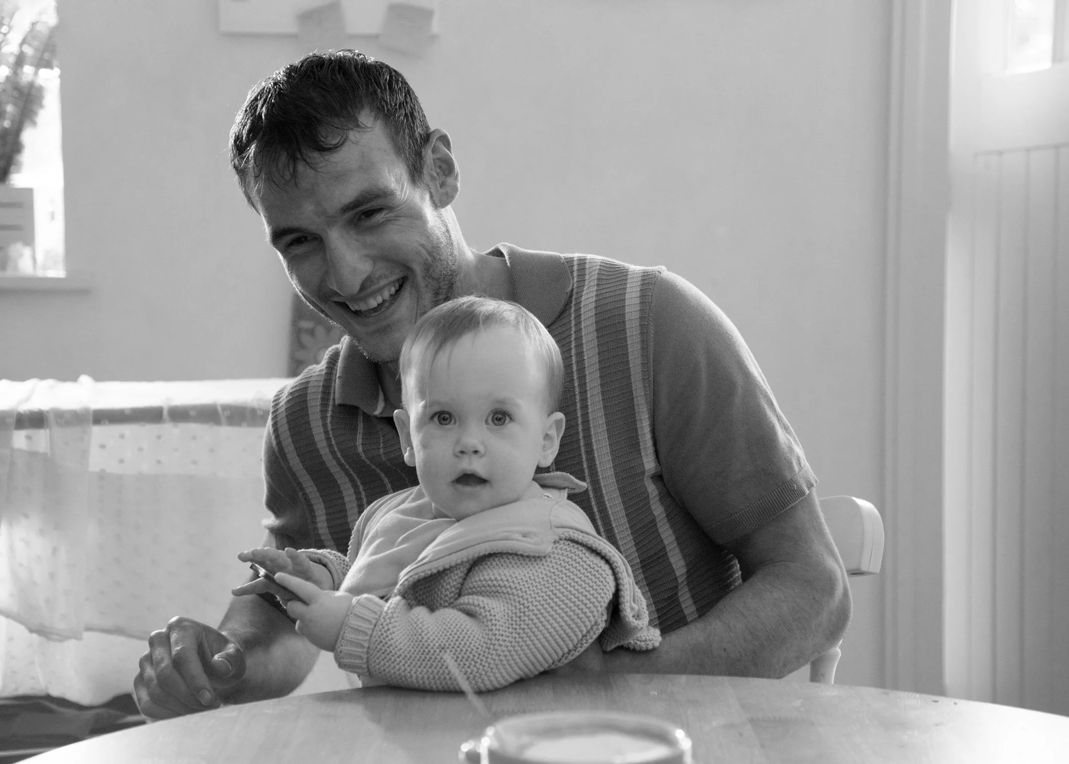 Behind the scenes photo. A man with short hair and a beard smiling and leaning over a table, holding a small child with light-colored hair and wide eyes, who is looking straight at the camera. The background shows a window and a wall.