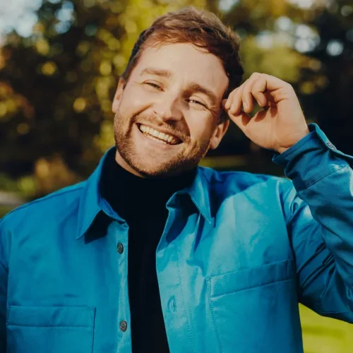 Smiling man with short brown hair and beard wearing a blue jacket and black shirt outdoors with trees in the background.