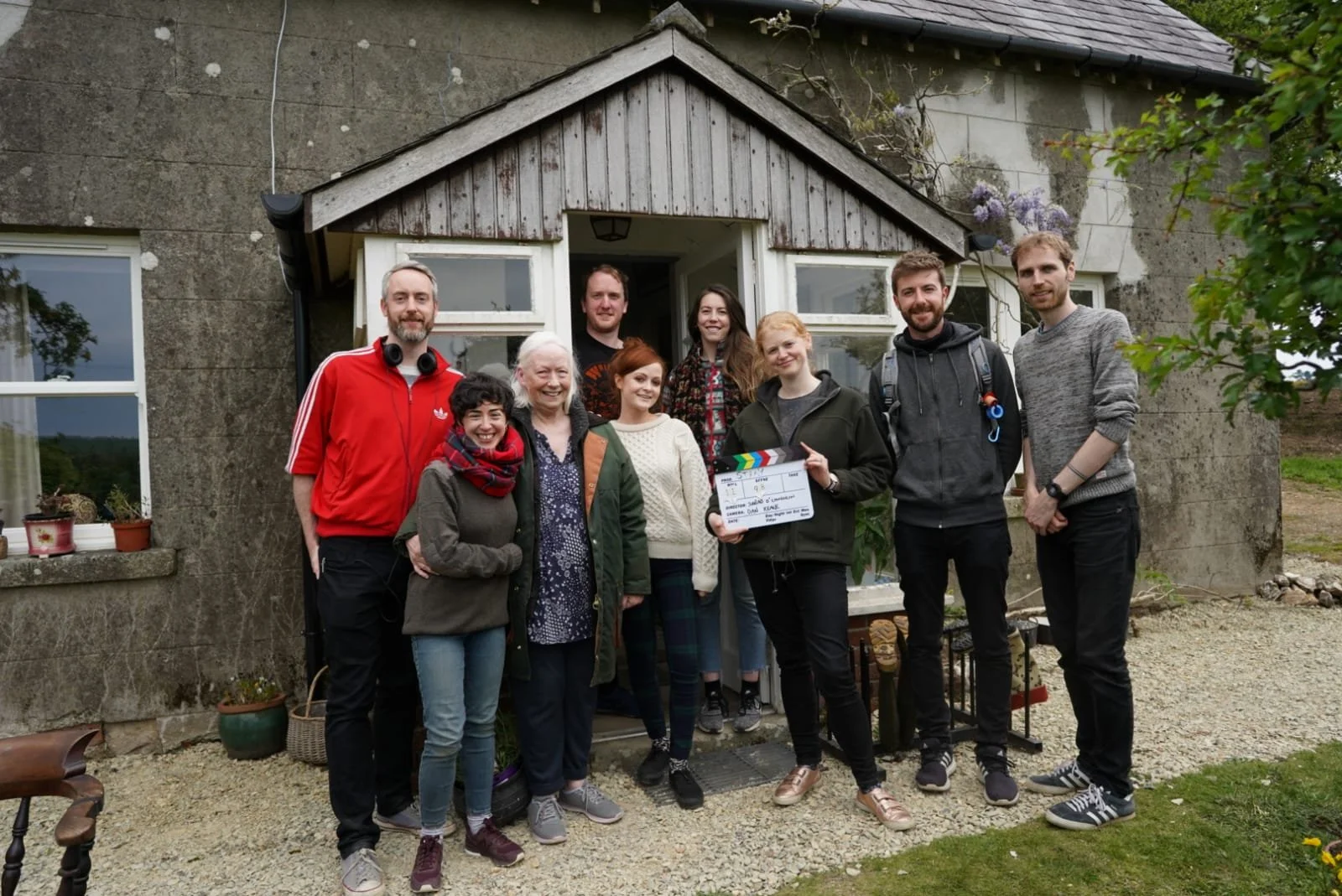 A group of ten people standing in front of a house, smiling and holding a clapperboard, with some potted plants on the windowsill and outdoor greenery around.
