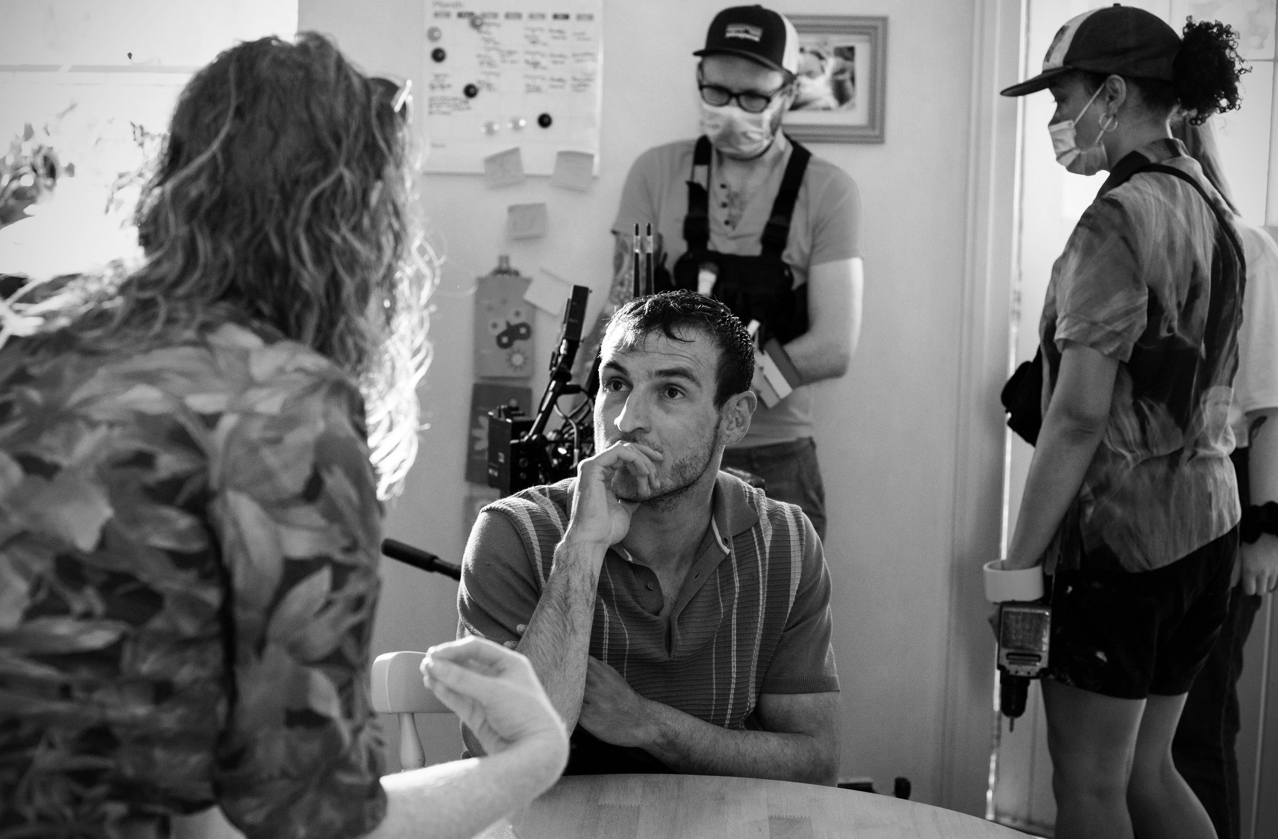 A man is seated at a table, resting his chin on his hand, looking thoughtfully at a woman with curly hair who is facing away from the camera. Three women in the background are wearing masks and gloves, in a clinical or laboratory setting.