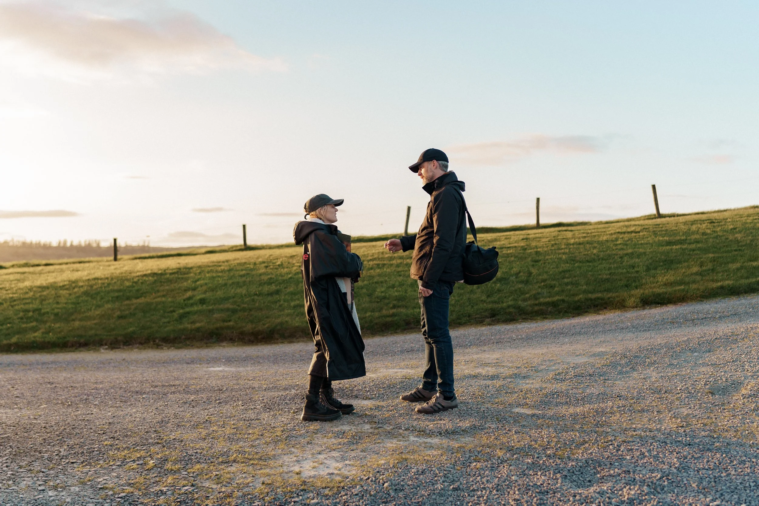 A man and a woman standing outdoors on a gravel path, having a conversation during sunset, both wearing black jackets and caps, with the woman holding a drink and the man carrying a black shoulder bag, with green grass and a fence in the background.