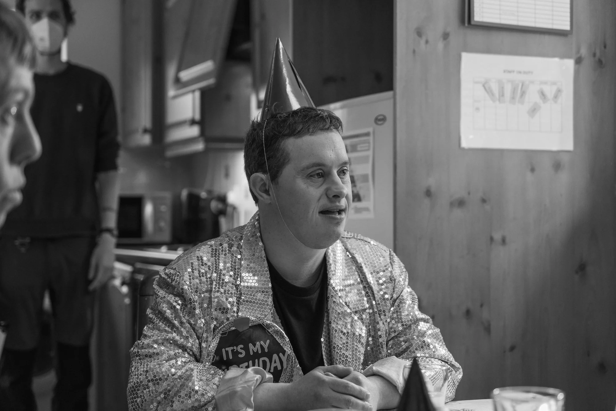 Behind the scenes photo from a film set. Person wearing a shiny, sequin-covered jacket and a birthday badge with the message "It's my birthday" sitting at a table during an celebration event.
