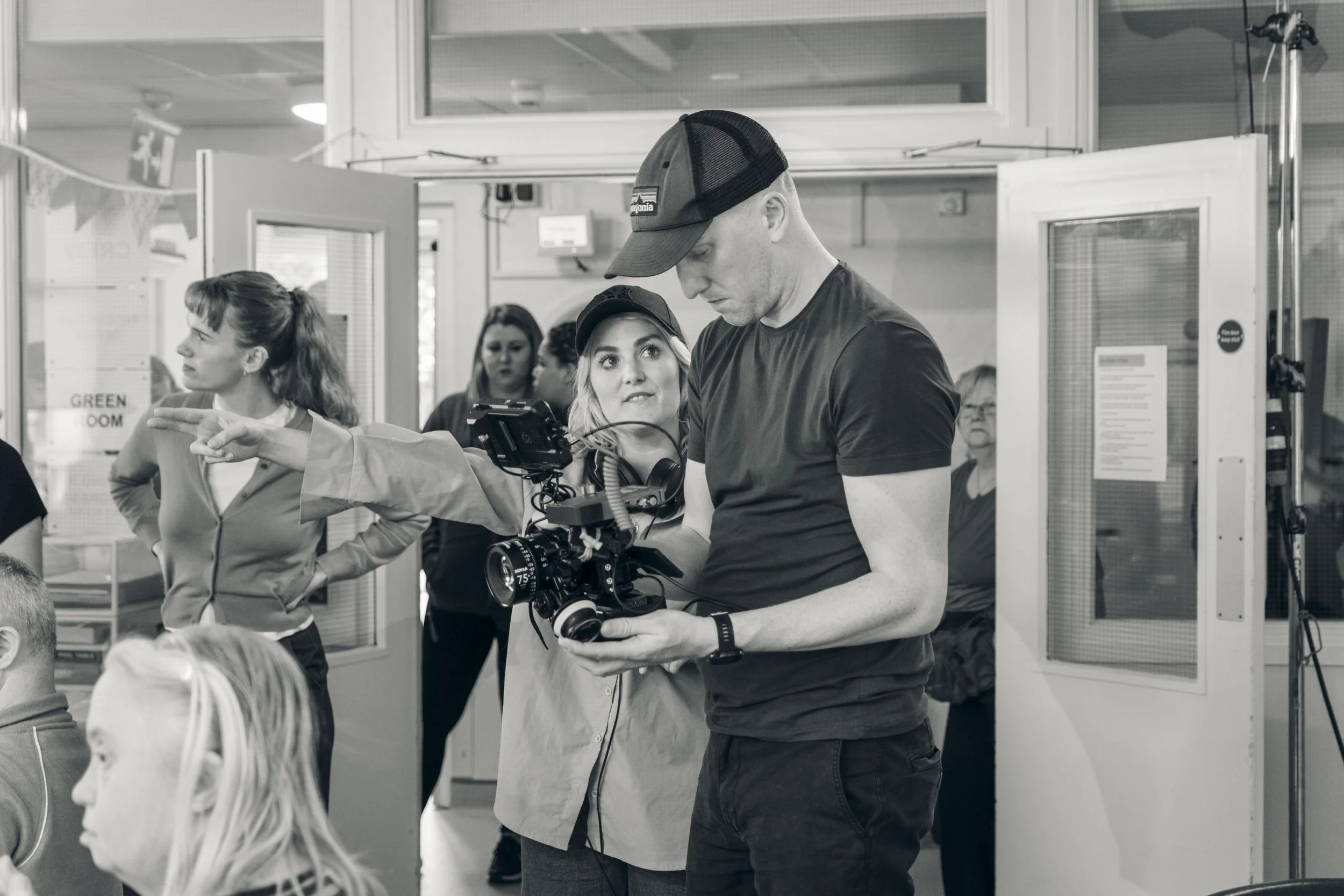 A young woman is assisting a man operating a professional camera, likely on a film or television set. The scene is inside a building with several people in the background, some looking towards the camera. The setting suggests a production environment