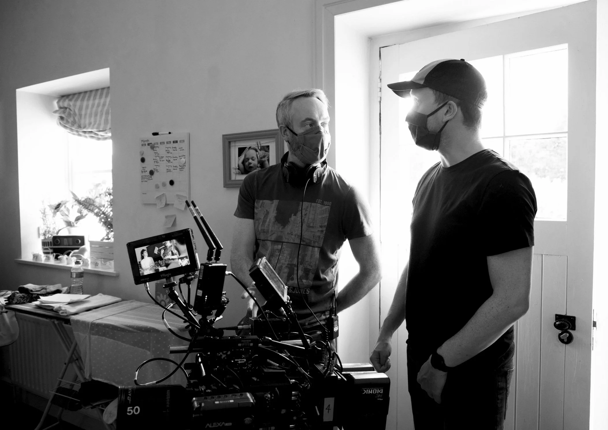 Two men wearing face masks shaking hands in a room with a window, a photograph on the wall, and various equipment including a camera and monitor on a table.