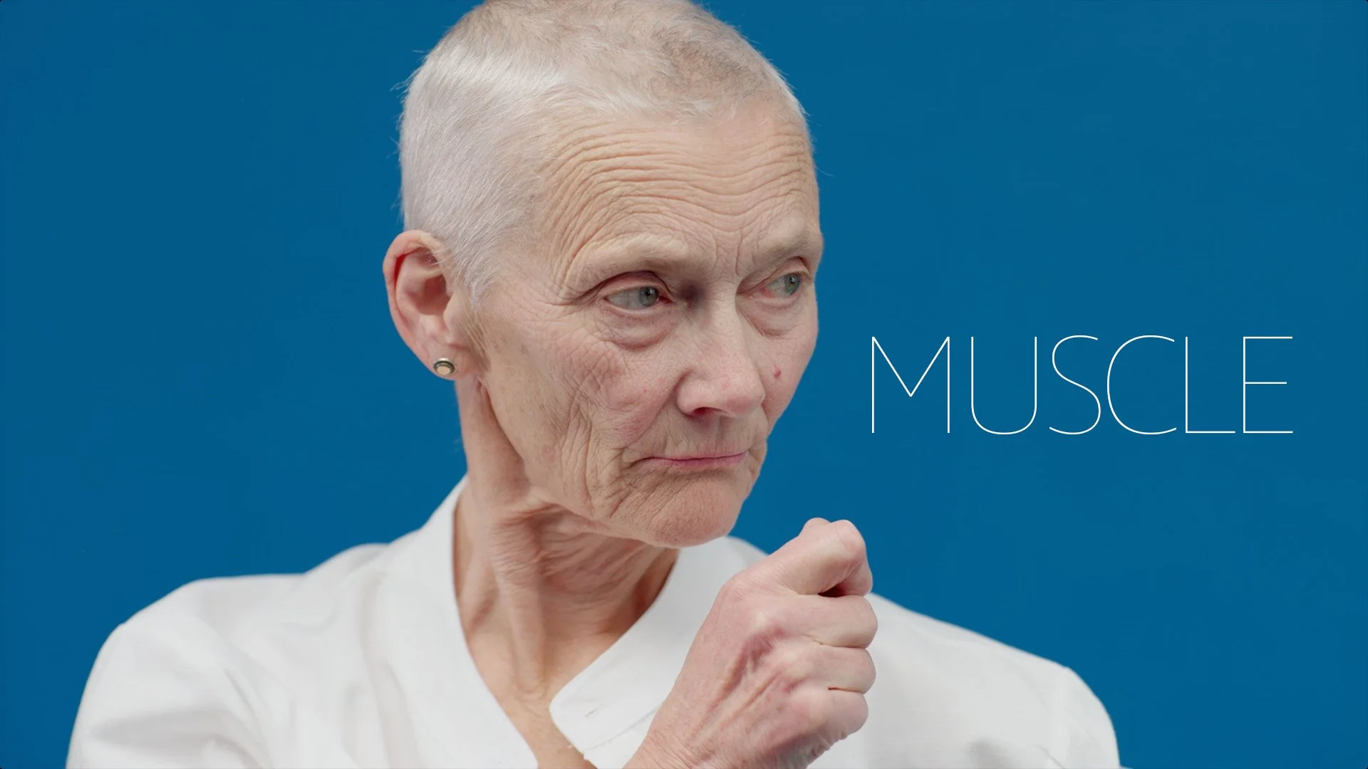 An elderly woman with short gray hair and earrings is looking to her right, with a serious expression. She is wearing a white shirt and has her right hand raised near her chin. The background is solid blue, and the word 'MUSCLE' is written in large t