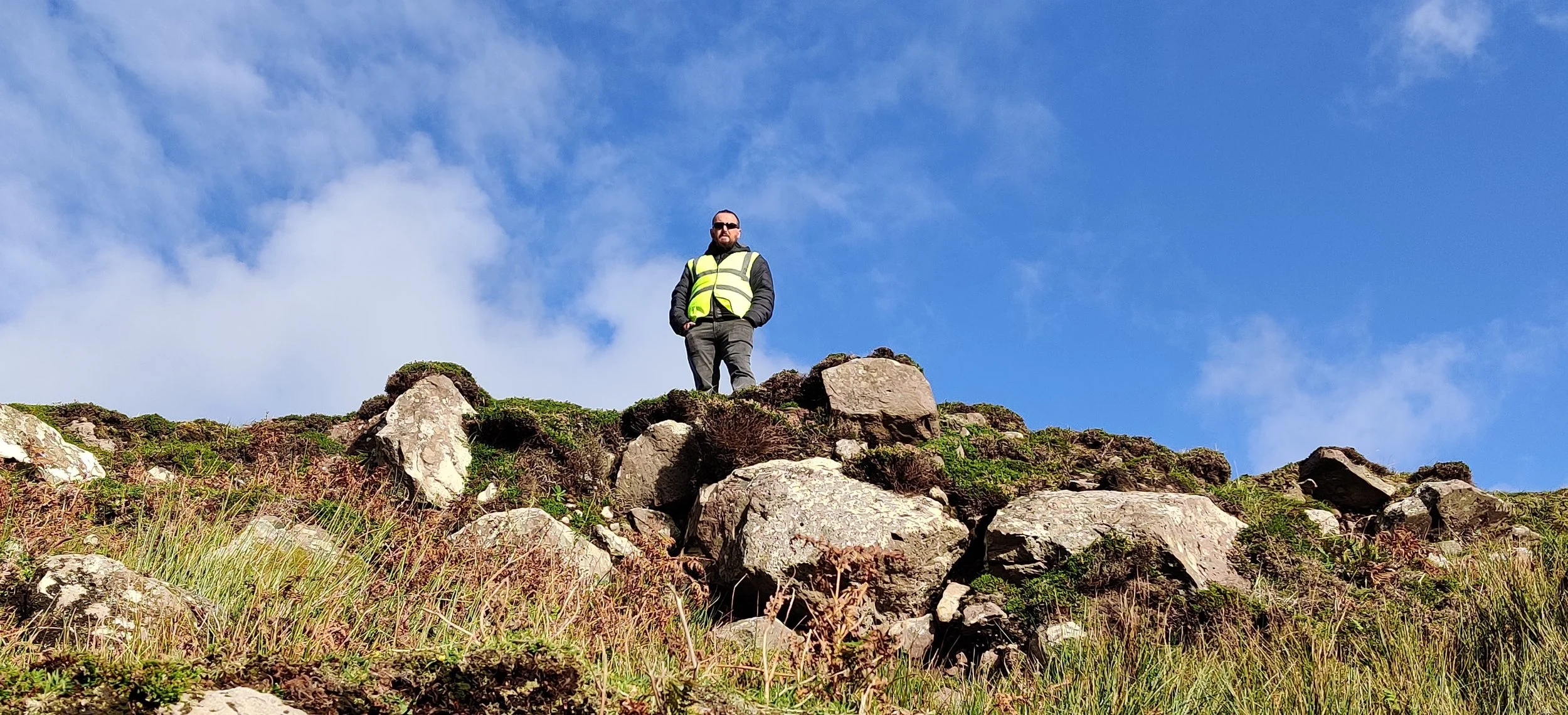 A man standing on top of a rocky hill with a blue sky and clouds in the background. He is wearing sunglasses, a black jacket, gray pants, and a yellow safety vest.