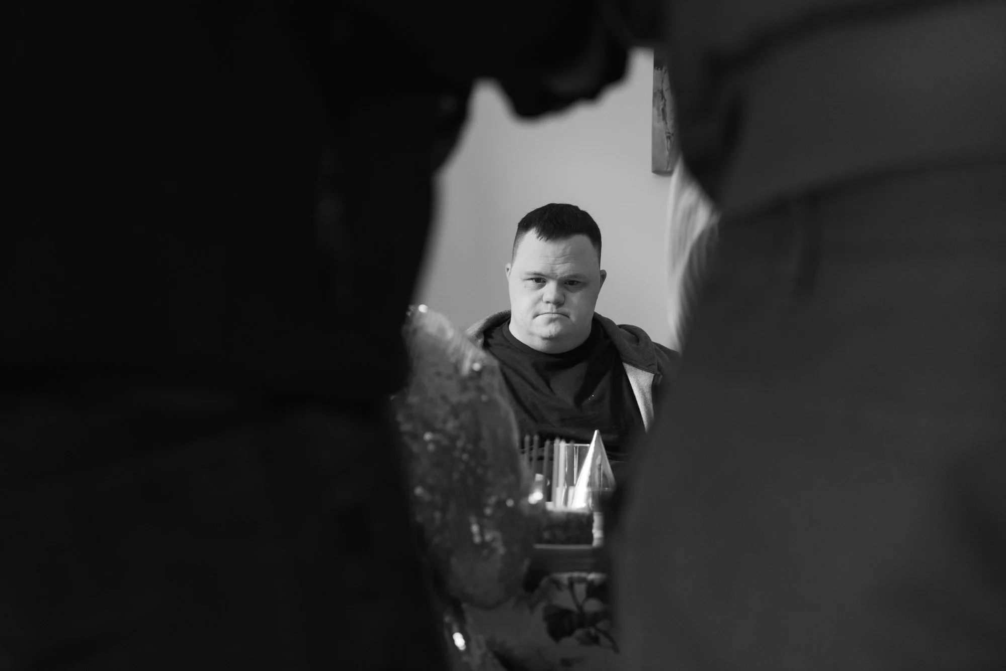 Behind the scenes photo from a film set. A young man with short dark hair and a serious expression, sitting at a table with multiple glasses and bottles, surrounded by others in a dim indoor setting.