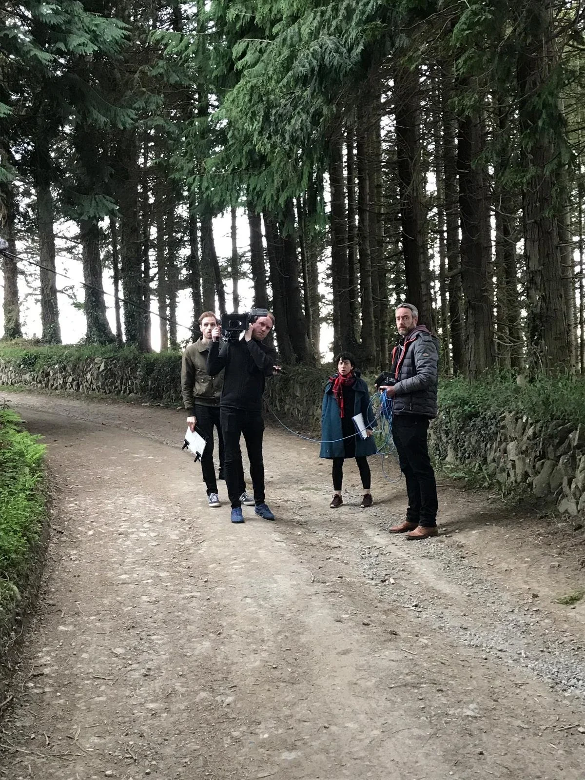 Group of four people standing on a dirt path in a wooded area, engaged in filming or photography with equipment.