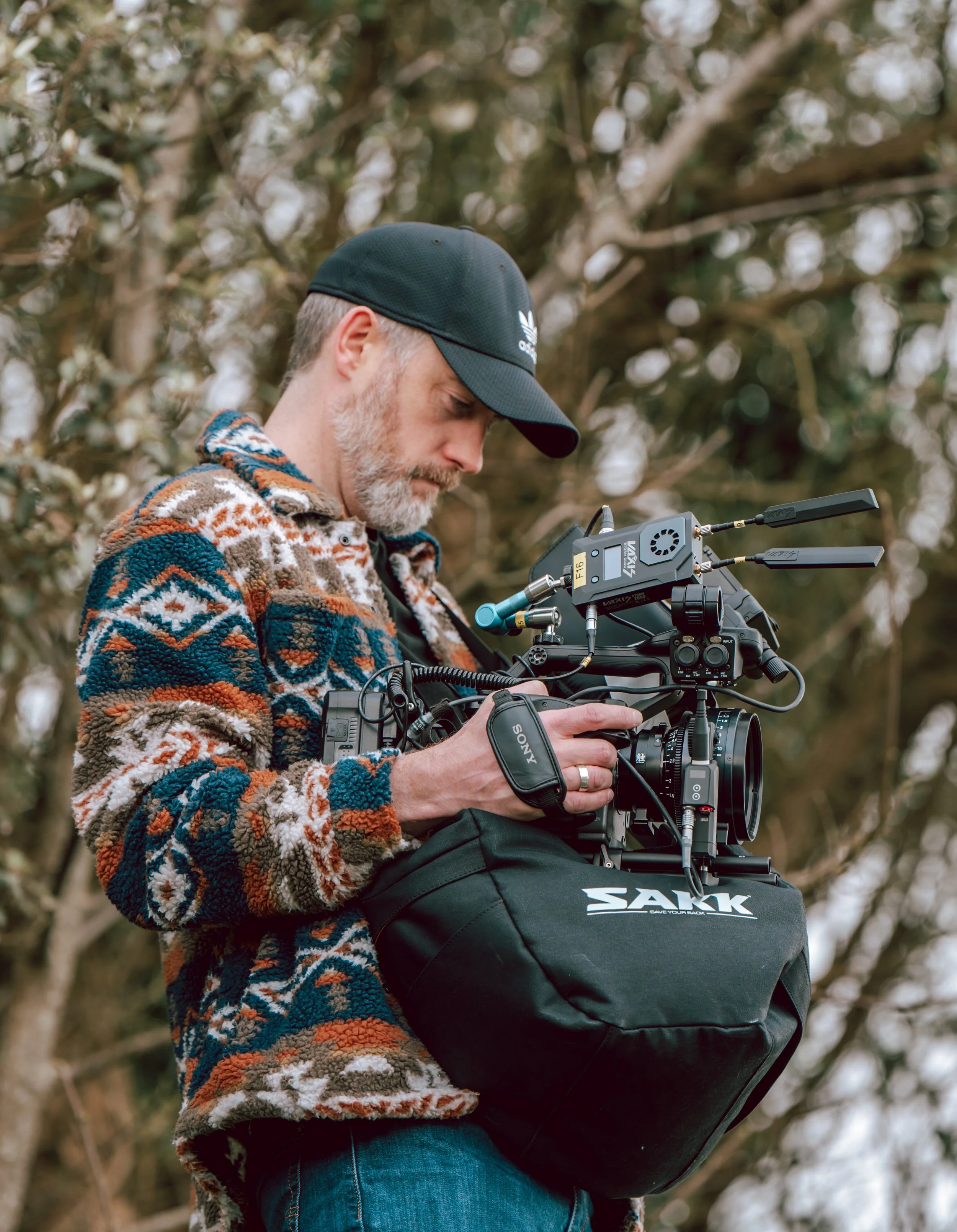 A man with a gray beard and a black cap operating a professional camera outdoors, surrounded by trees.