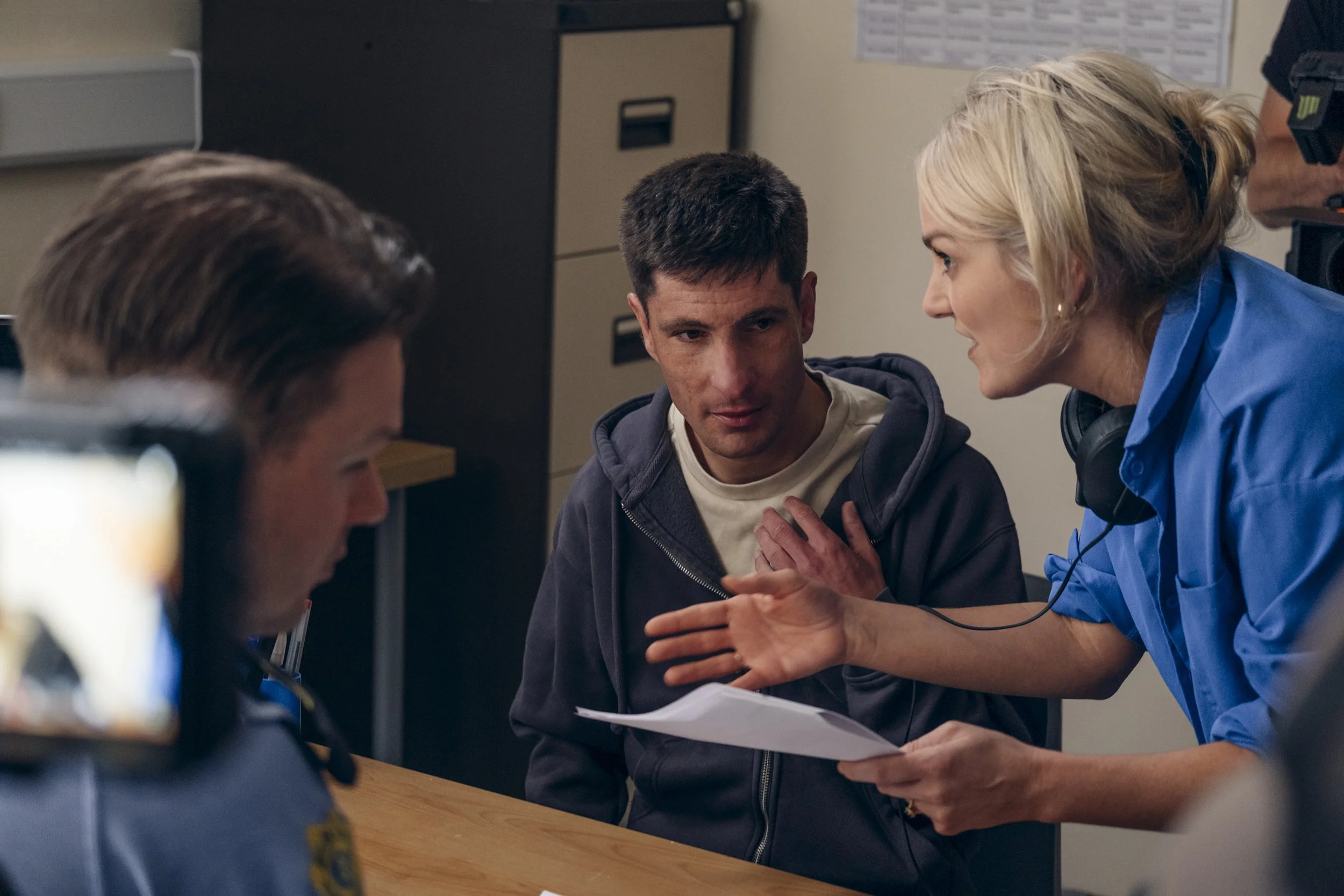 A woman in a blue shirt and headphones is talking to a young man wearing a hoodie, sitting at a table with papers in front of them. They appear to be in a clinical or office setting, engaged in a conversation.